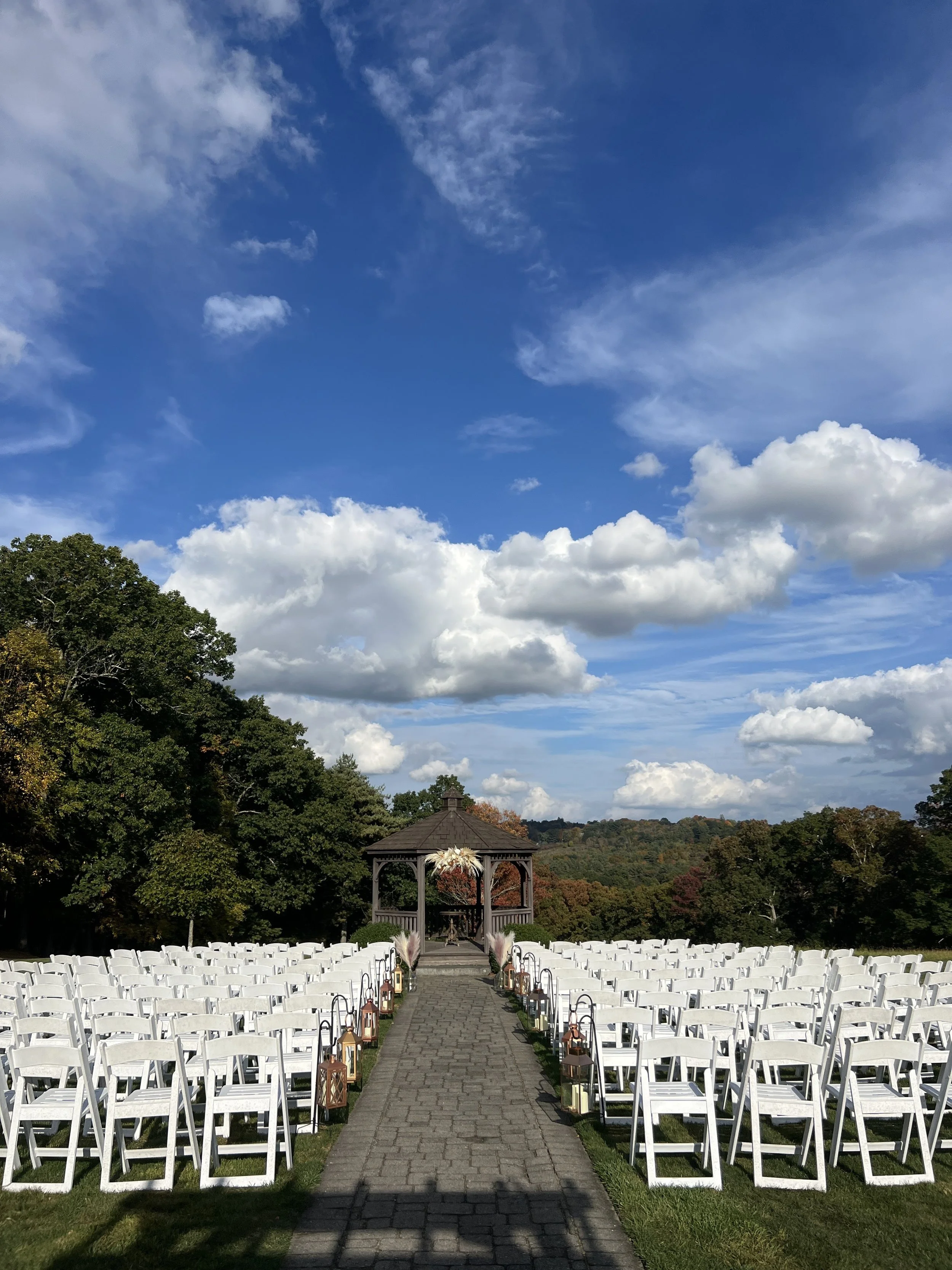 Outdoor wedding setup with a path leading to a gazebo, white chairs arranged on either side, and lanterns along the aisle, under a partly cloudy sky with trees in the background.
