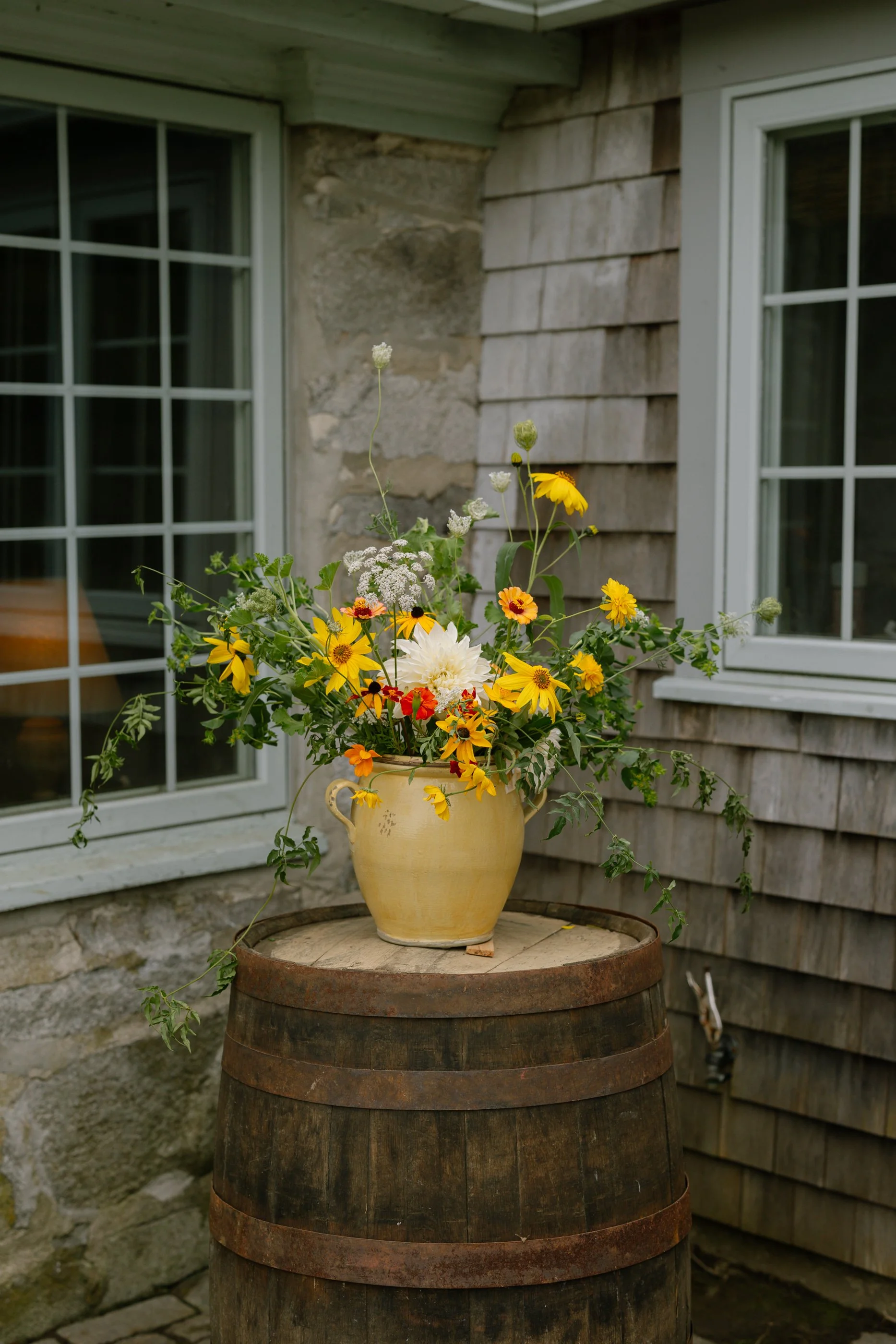 A yellow ceramic vase filled with colorful wildflowers, placed on a wooden barrel outside a house with stone and shingle siding.