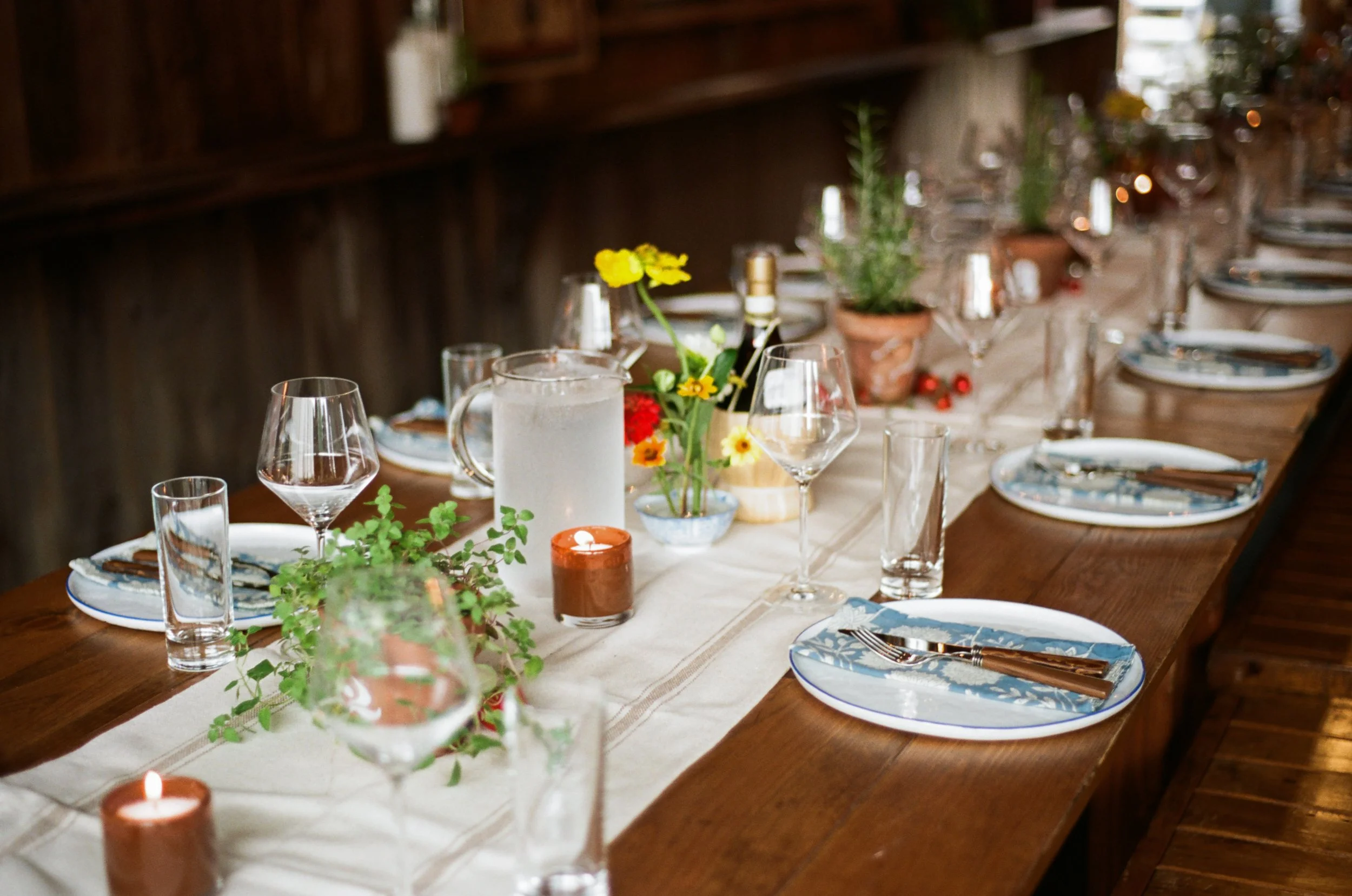 A rustic wooden dining table set for a gathering with plates, glasses, and silverware, decorated with potted plants, flowers, and candles.