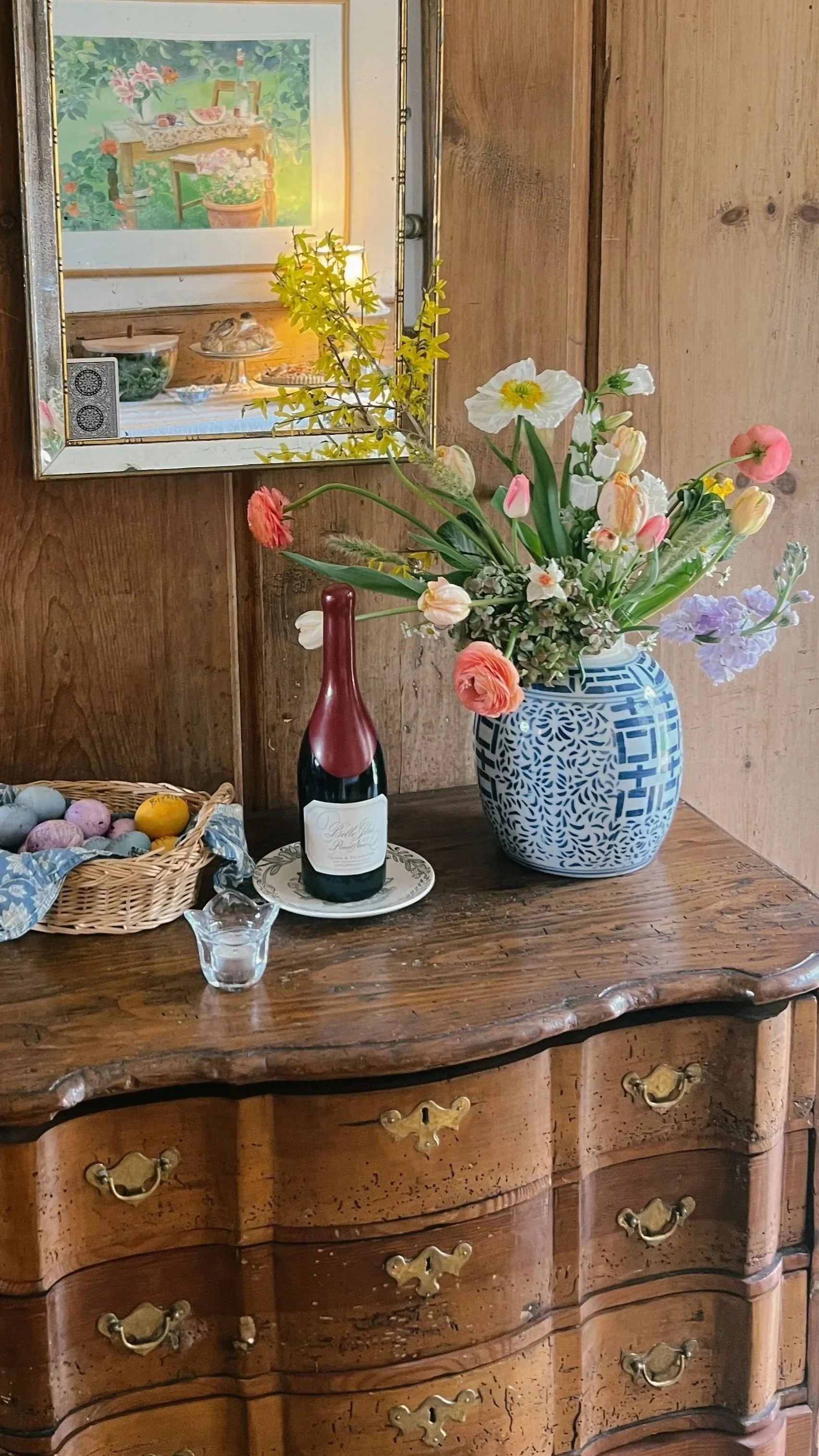 A wooden dresser with brass handles decorated with a large bouquet of assorted flowers in a blue and white patterned vase, a bottle of sparkling wine on a saucer, a small glass pitcher, and a basket of painted Easter eggs. A mirror above reflects a s
