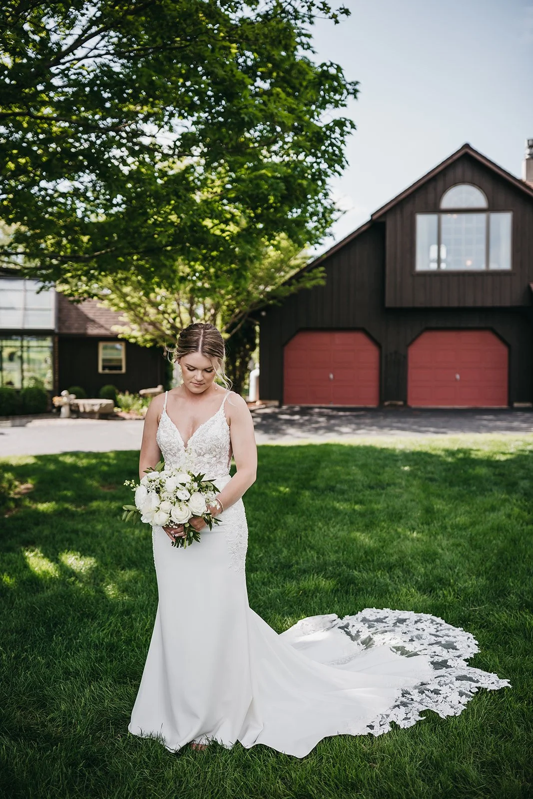 Bride in a white lace wedding dress holding a bouquet of white flowers in a backyard with green grass and a large tree, with a dark-colored house and red garage doors in the background.