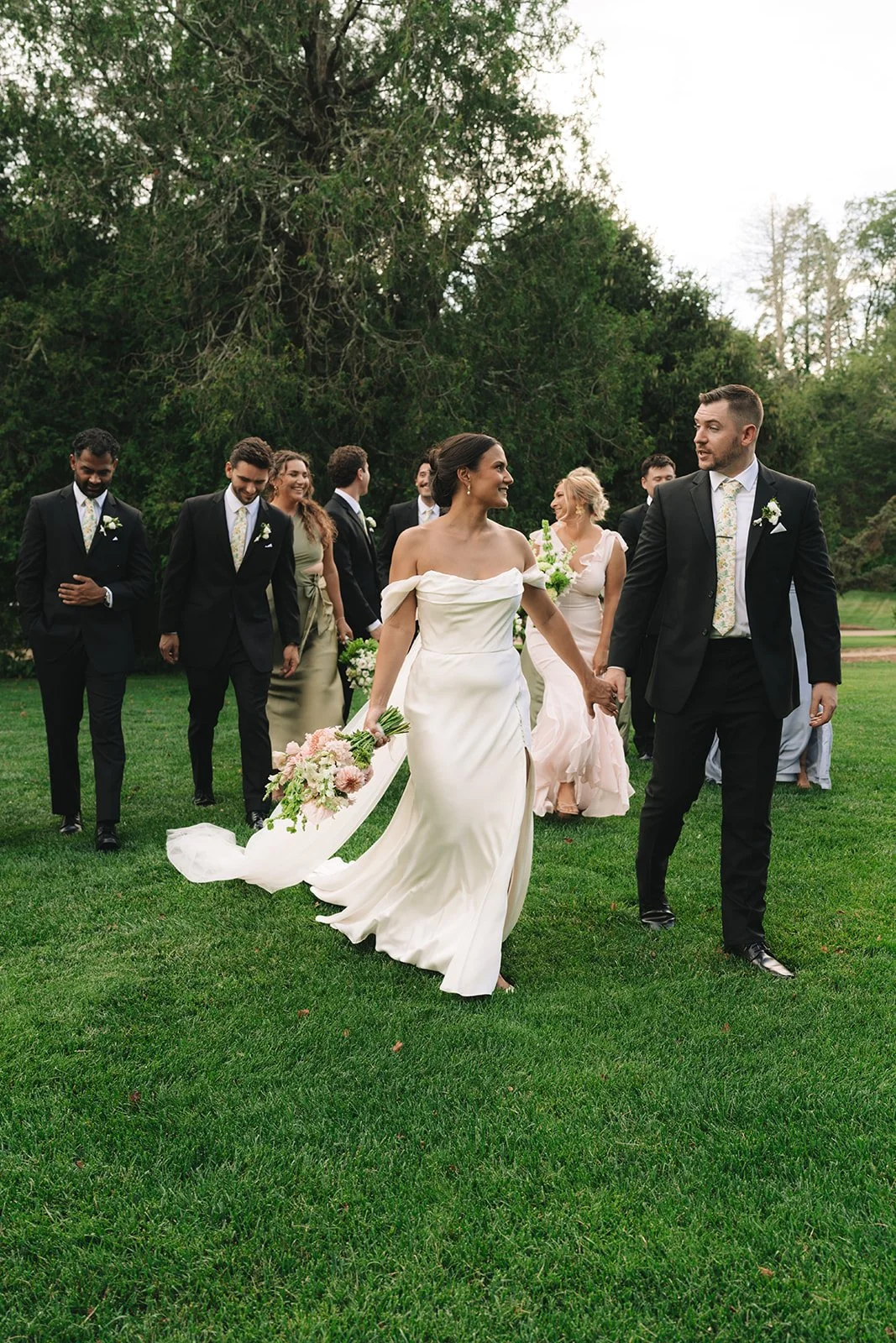 Bride and groom holding hands walking on a grassy area with wedding party in the background at an outdoor wedding.