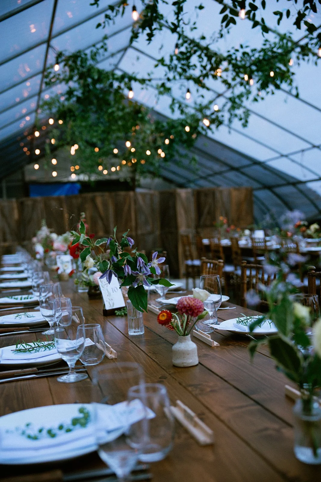 A decorated dinner table with floral arrangements in a greenhouse or similar setting, with string lights hanging from the ceiling.