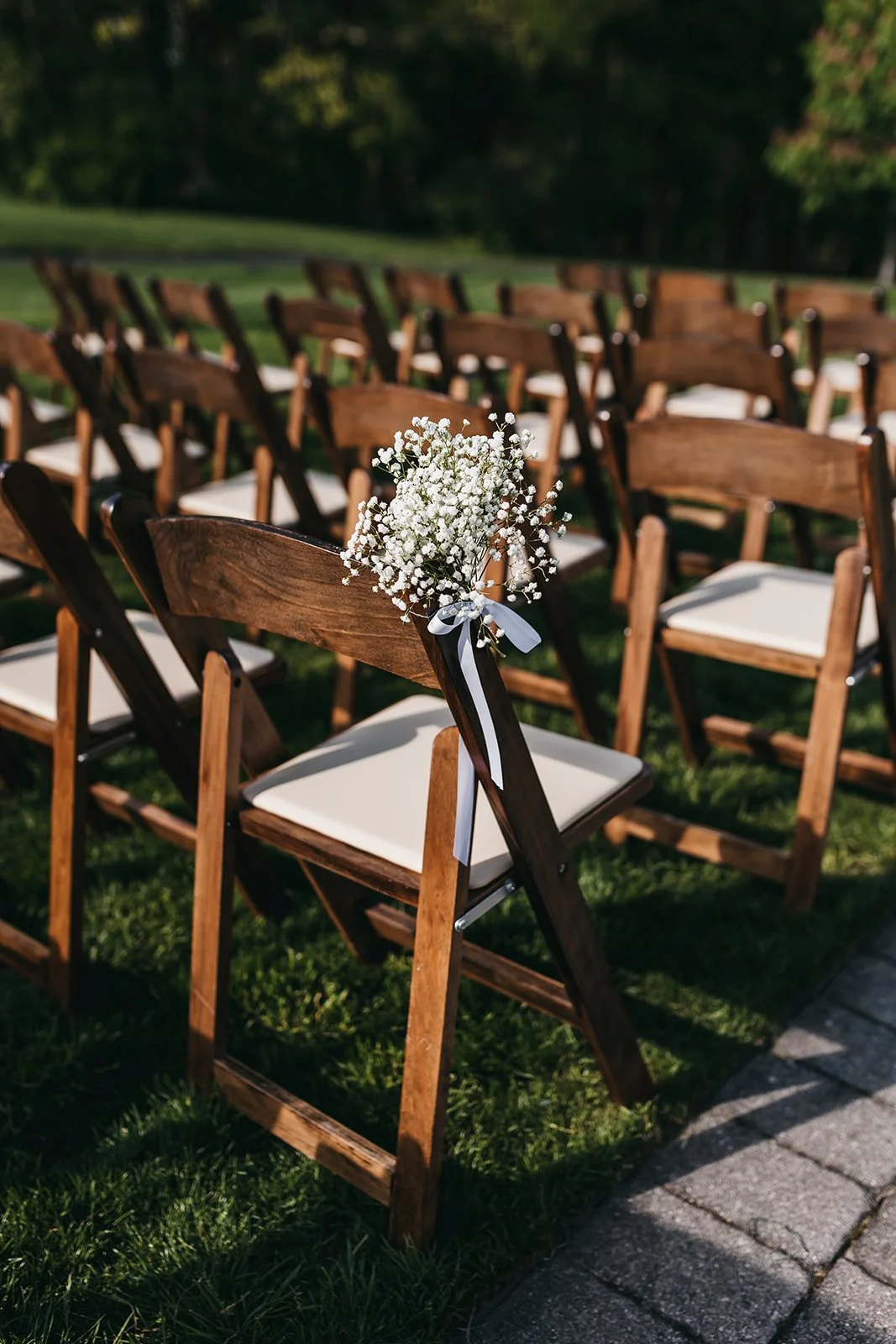Wooden chairs with white cushions arranged outdoors, with a small bouquet of white baby's breath flowers attached to one chair, indicating a wedding ceremony setting.