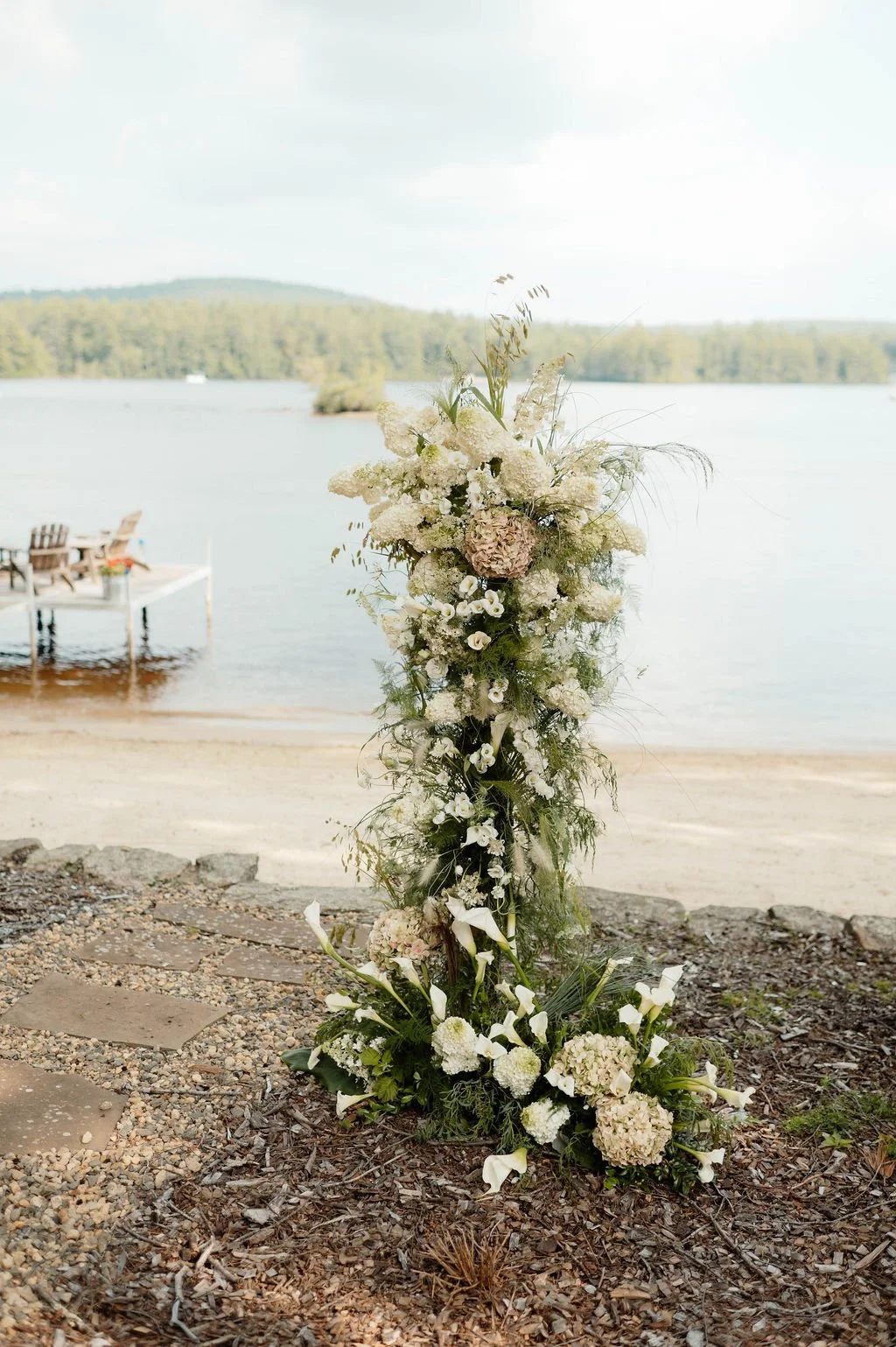 A floral arrangement with white and pale pink flowers, greenery, and calla lilies near a lakeside, with a dock and chairs in the background.