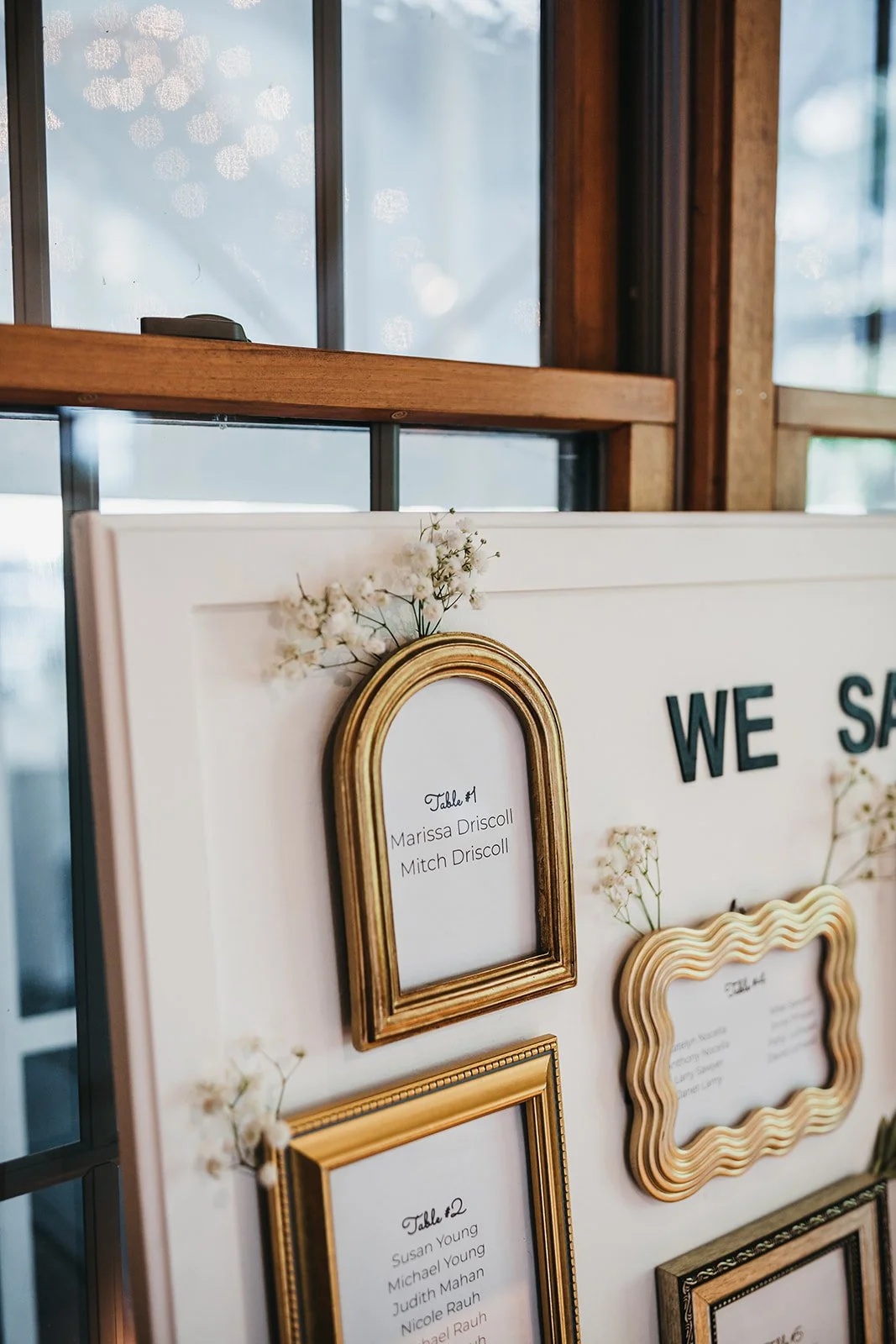Wedding seating chart with gold frames and small white flowers, featuring tables and guest names, displayed on a white board near windows.