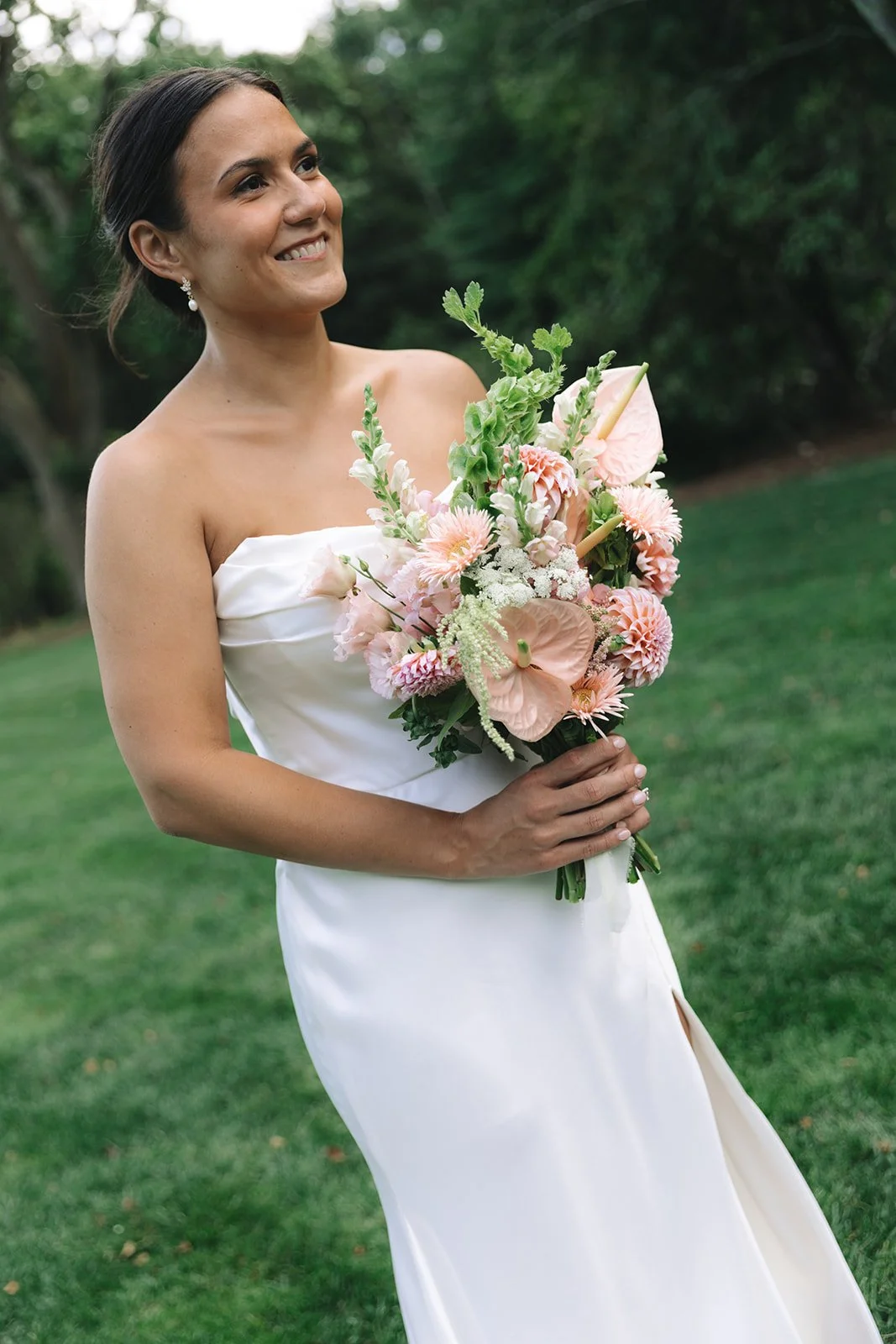 A woman in a white strapless dress holding a bouquet of pink and white flowers outdoors with green grass and trees in the background.