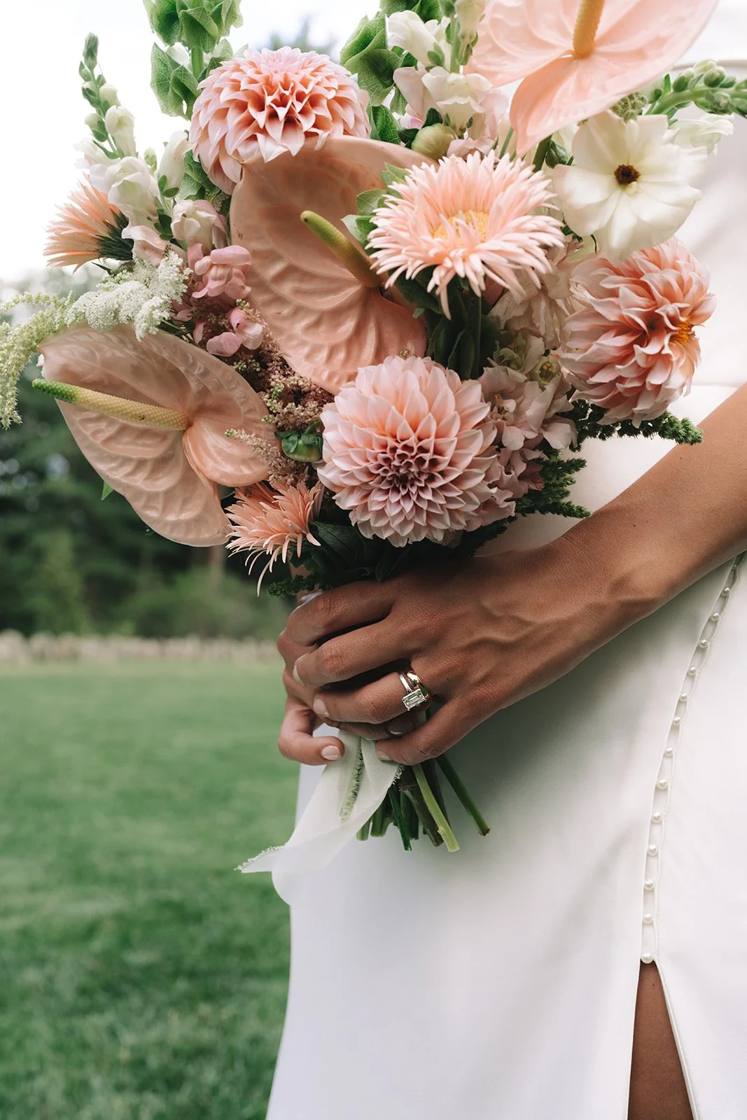 Close-up of a bride holding a bouquet of pink and white flowers, including dahlias, anthuriums, and anemones, with a wedding ring on her finger and a white dress with pearl buttons.