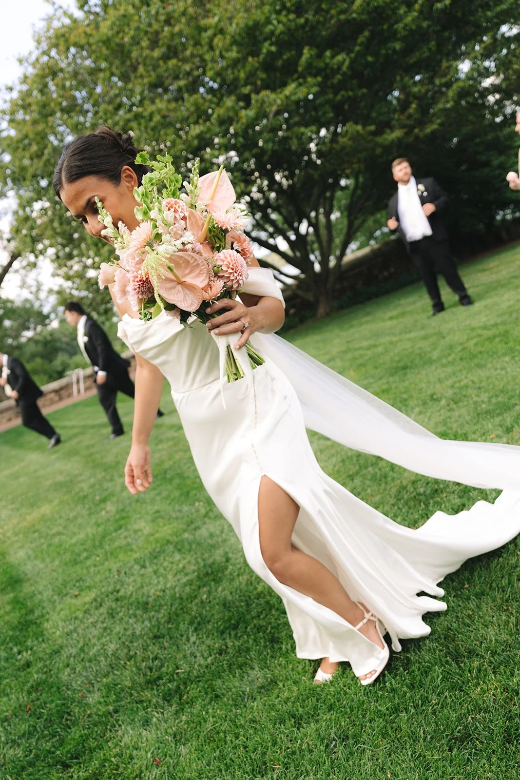 A bride in a white wedding dress holding a bouquet of pink flowers, walking on grass during an outdoor wedding ceremony, with groomsmen standing in the background under large trees.