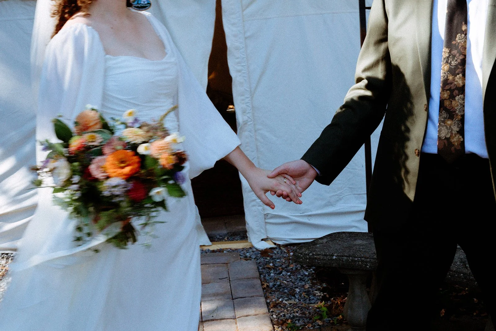 A bride and groom holding hands during their wedding ceremony. The bride is holding a colorful bouquet of flowers, and they are standing outdoors near a white tent.