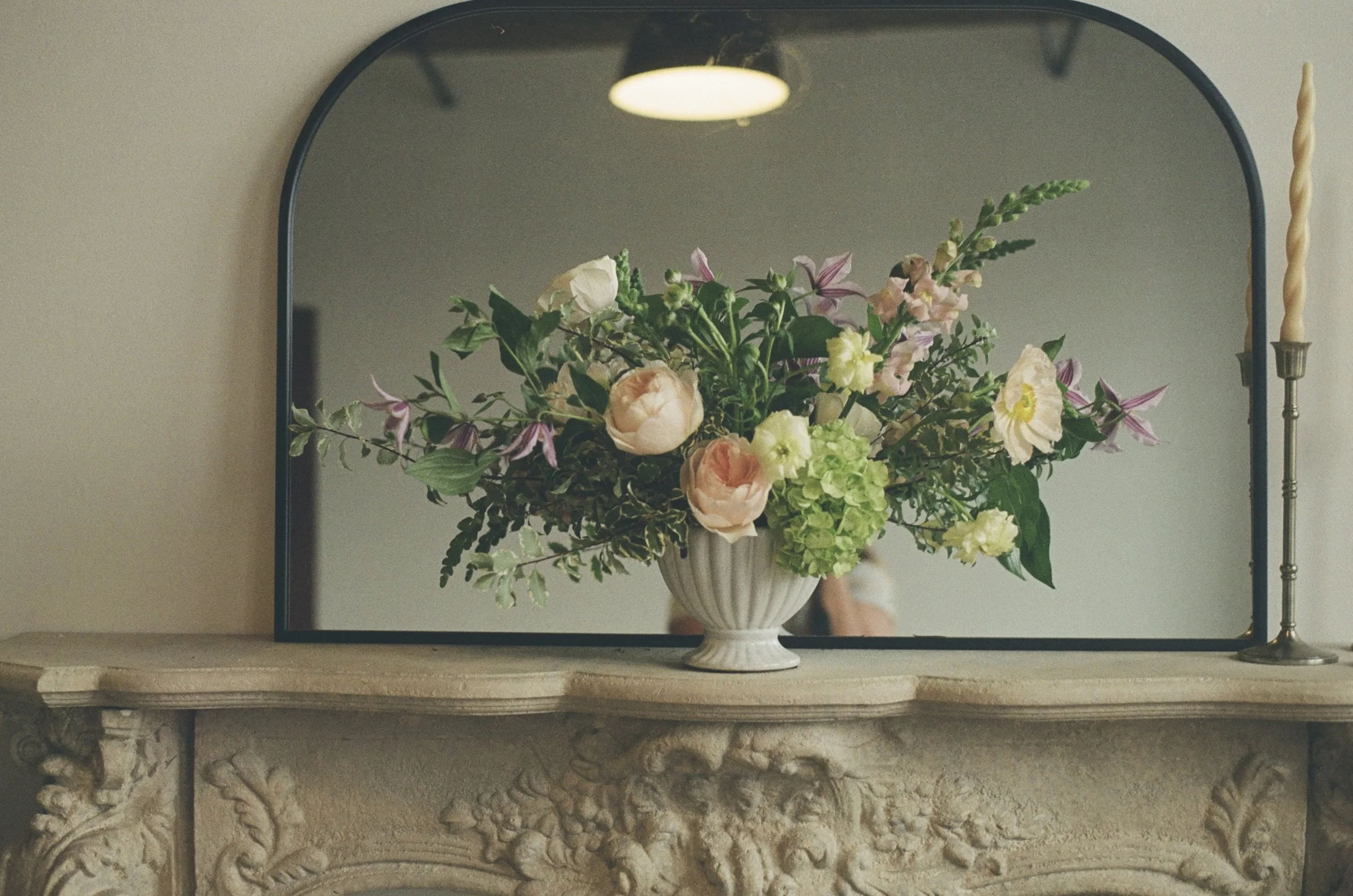 A floral arrangement in a white vase reflected in a mirror on a decorative fireplace mantel, with a candlestick to the right and a ceiling light visible in the reflection.