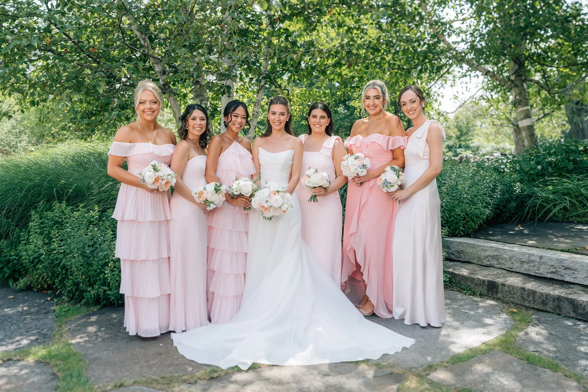 A group of eight women, including a bride and bridesmaids, standing outdoors under trees, dressed in elegant pink and white dresses, holding bouquets of flowers, smiling for a photo.