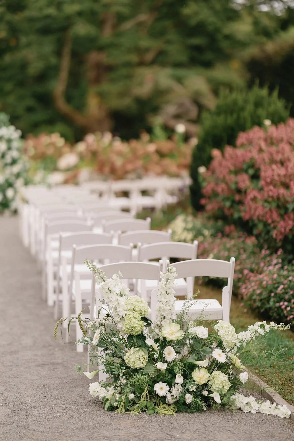 A white floral arrangement with white and cream flowers and greenery at the beginning of a row of white chairs, set outdoors on a gravel path with pink and white flowering bushes and trees in the background, awaiting a wedding ceremony.