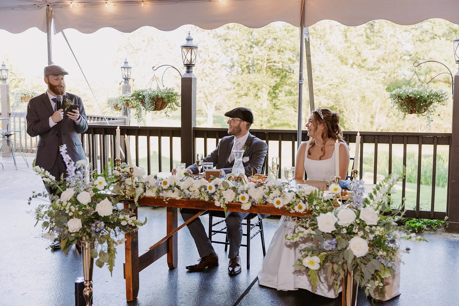 Wedding reception with bride and groom sitting at a decorated table, officiant speaking with a microphone, outdoors with trees in the background, floral arrangements on the table and hanging baskets.