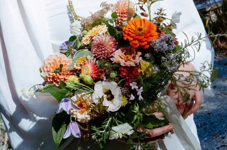 A bouquet of orange roses, white lilies, small white flowers, green leaves, and a dandelion seed head on a wooden surface.