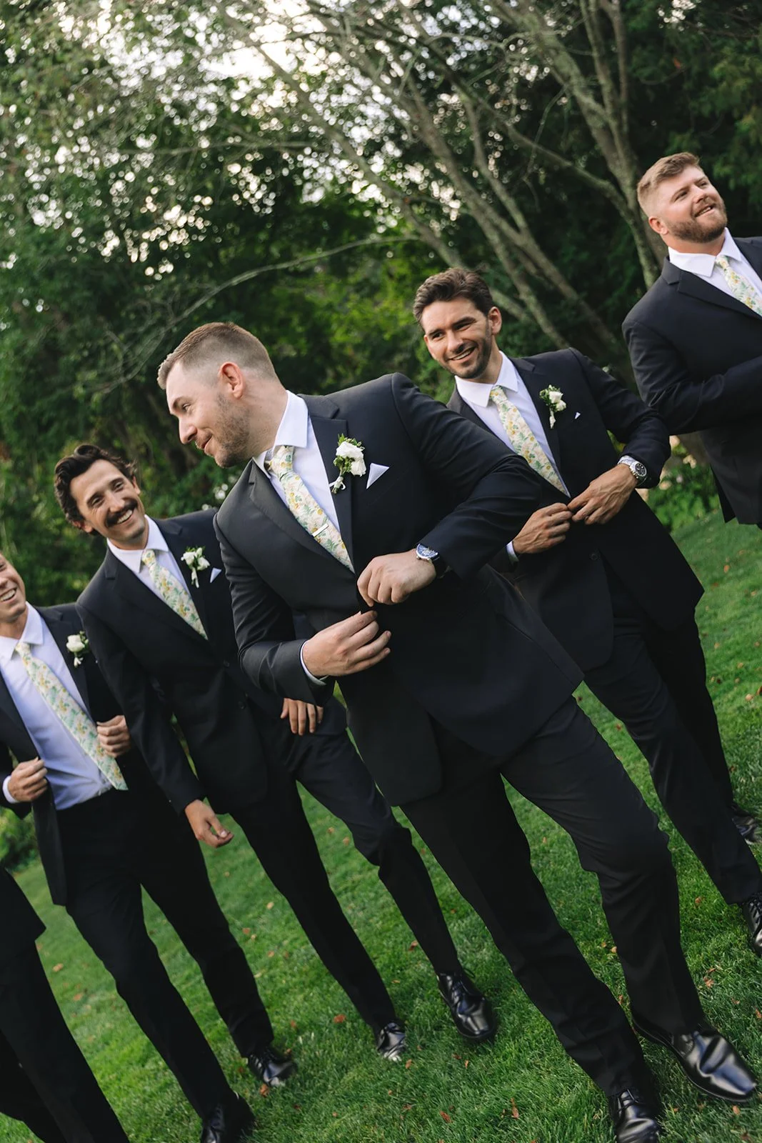 Group of men in black suits with floral ties and white boutonnieres standing outdoors on grass, laughing and chatting.