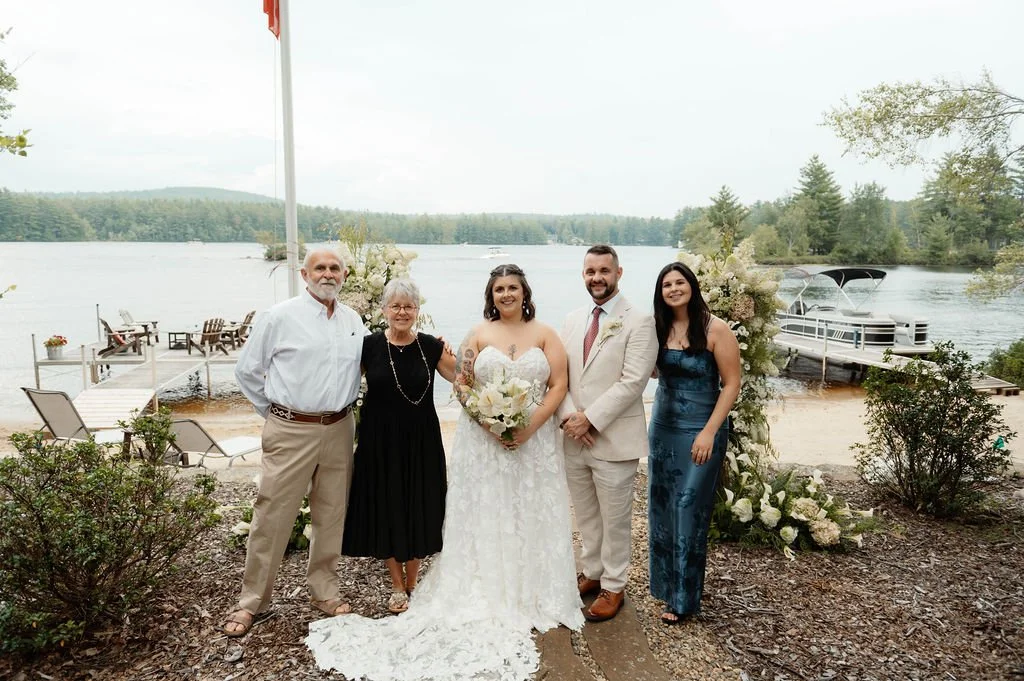 A wedding party stands outdoors by a lake, with five people including a bride in a white wedding dress holding a bouquet, a groom in a light suit, two women, and a man, surrounded by flowers and greenery.