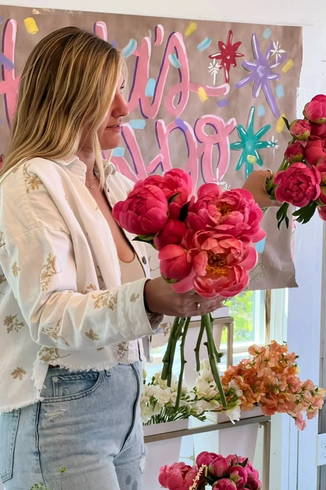 Woman arranging pink peony flowers at a celebration event with a colorful, handwritten 'HAPPY' sign in the background.