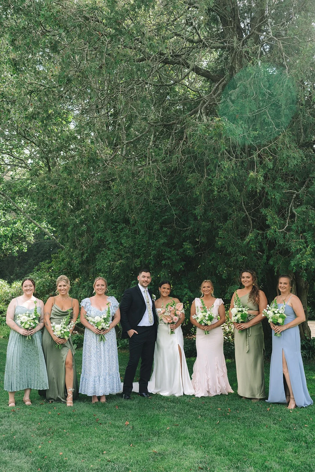 A wedding party poses outdoors in front of a large tree. The group includes the bride, groom, and six women in pastel-colored dresses, each holding a bouquet of flowers.