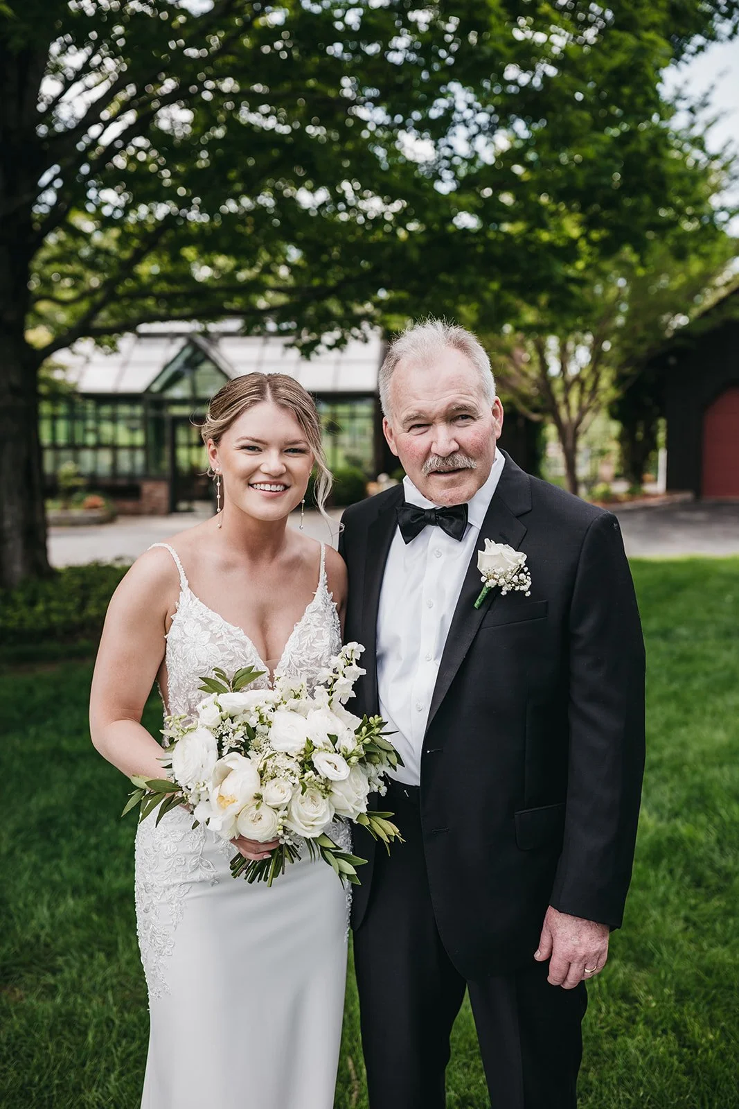 A bride in a white wedding dress holding a bouquet standing next to an older man in a black tuxedo with a black bow tie, outdoors with green trees and a greenhouse in the background.