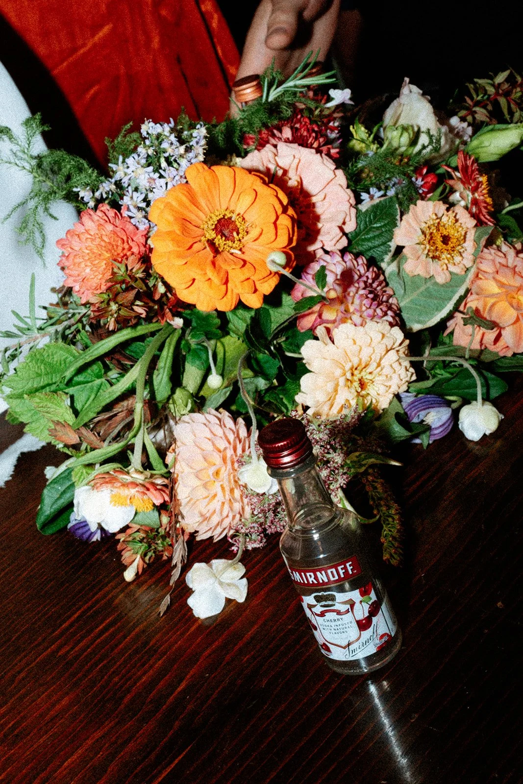 A bouquet of mixed flowers with various colors, including orange, pink, and white, resting on a dark wooden table next to a small bottle of Smirnoff cherry vodka.
