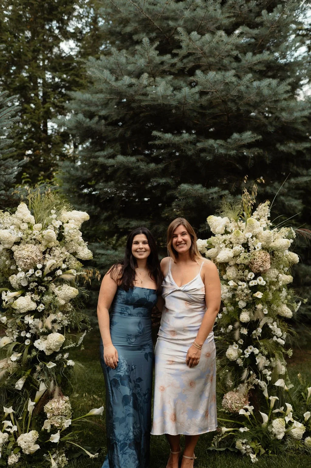 Two women standing outdoors between large floral arrangements of white flowers with greenery, in front of a large evergreen tree, smiling at the camera.