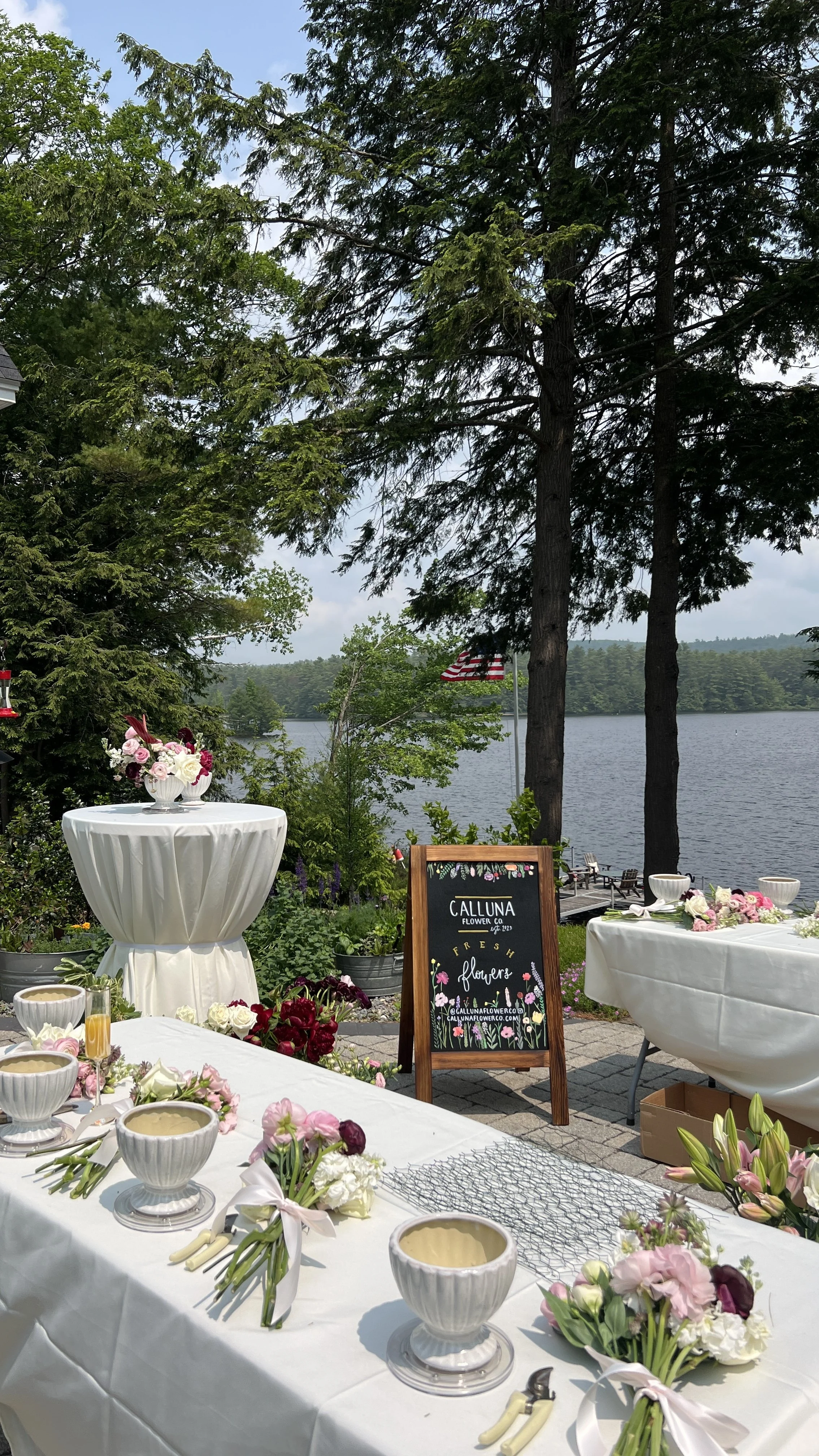 Decorated outdoor table set up by a lake with floral arrangements and a chalkboard sign promoting Calluna Flower Co., surrounded by trees and American flags.