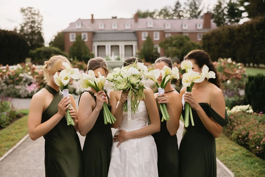 Group of five women in formal dresses holding bouquets of calla lilies, standing on a garden path in front of a large mansion.