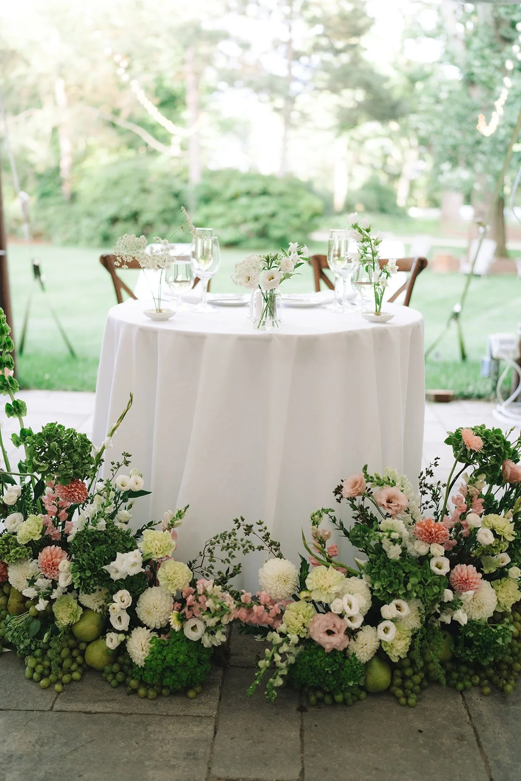 Round table with a white tablecloth, decorated with white and pale pink flowers in glass vases, set for a meal, surrounded by lush greenery and floral arrangements on the ground.