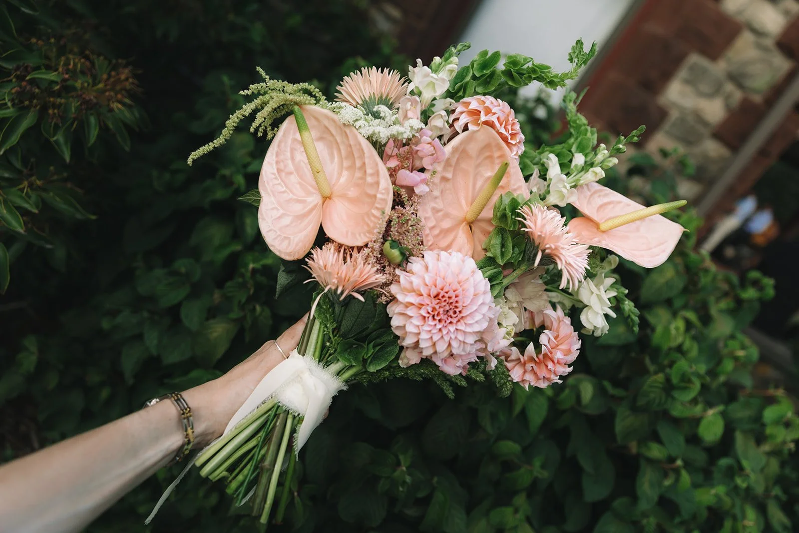 A person holding a bouquet of pink and white flowers including anthuriums, chrysanthemums, and other mixed blooms, with greenery, in an outdoor setting next to a brick wall.