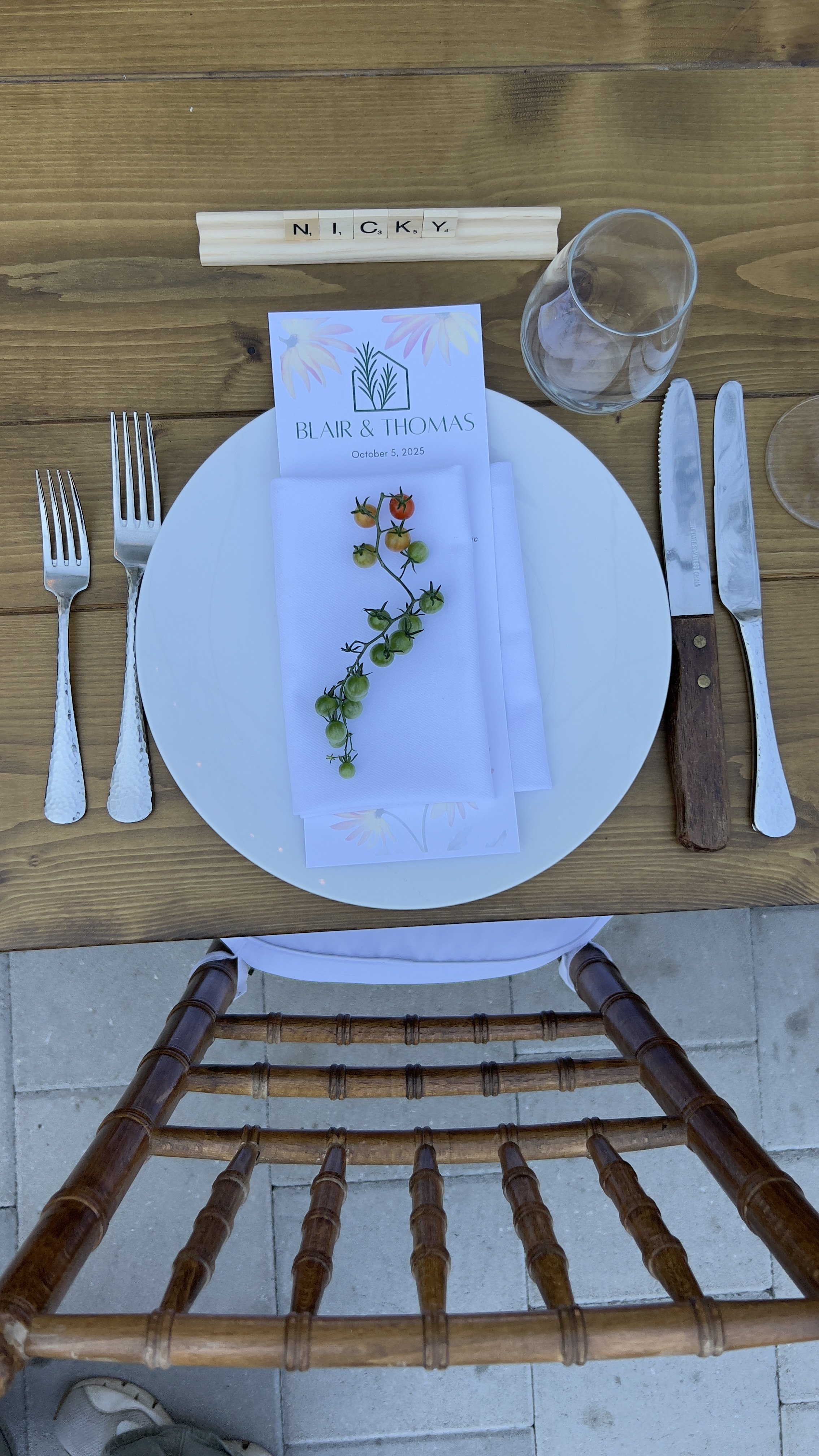 A wooden table set for a meal with a white plate, silverware, a folded napkin with a small tomato vine decoration, a clear wine glass, a small place card with the names Blair & Thomas and the date October 5, 2025, and Scrabble tiles spelling 'NICKY'.