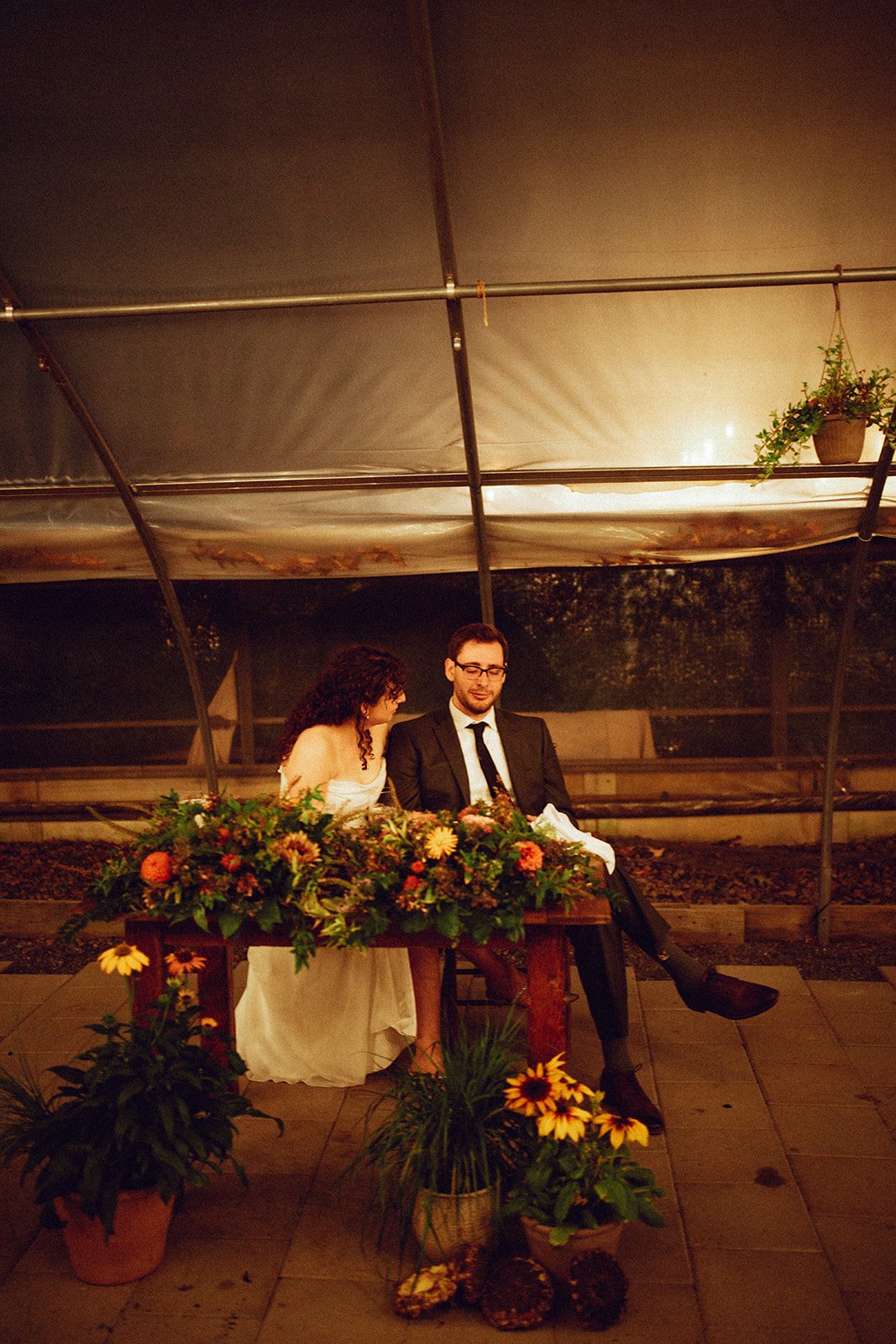 A couple sitting at a table decorated with a large floral arrangement in an outdoor greenhouse at night.