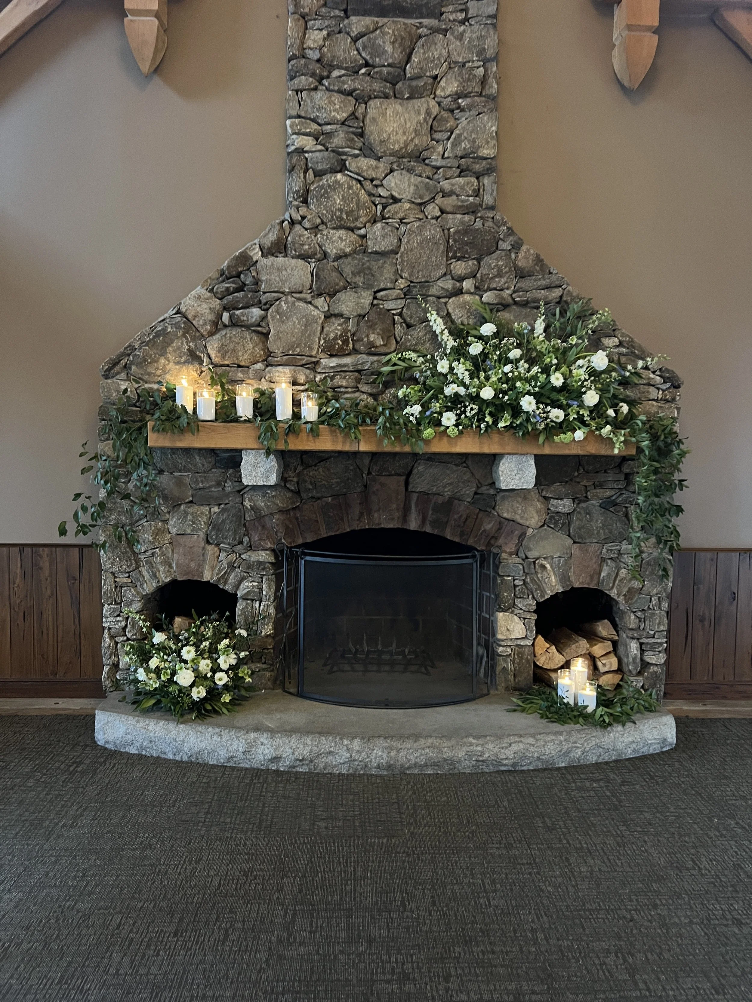 A rustic stone fireplace decorated with white flowers, candles, and green foliage for a celebration or event.
