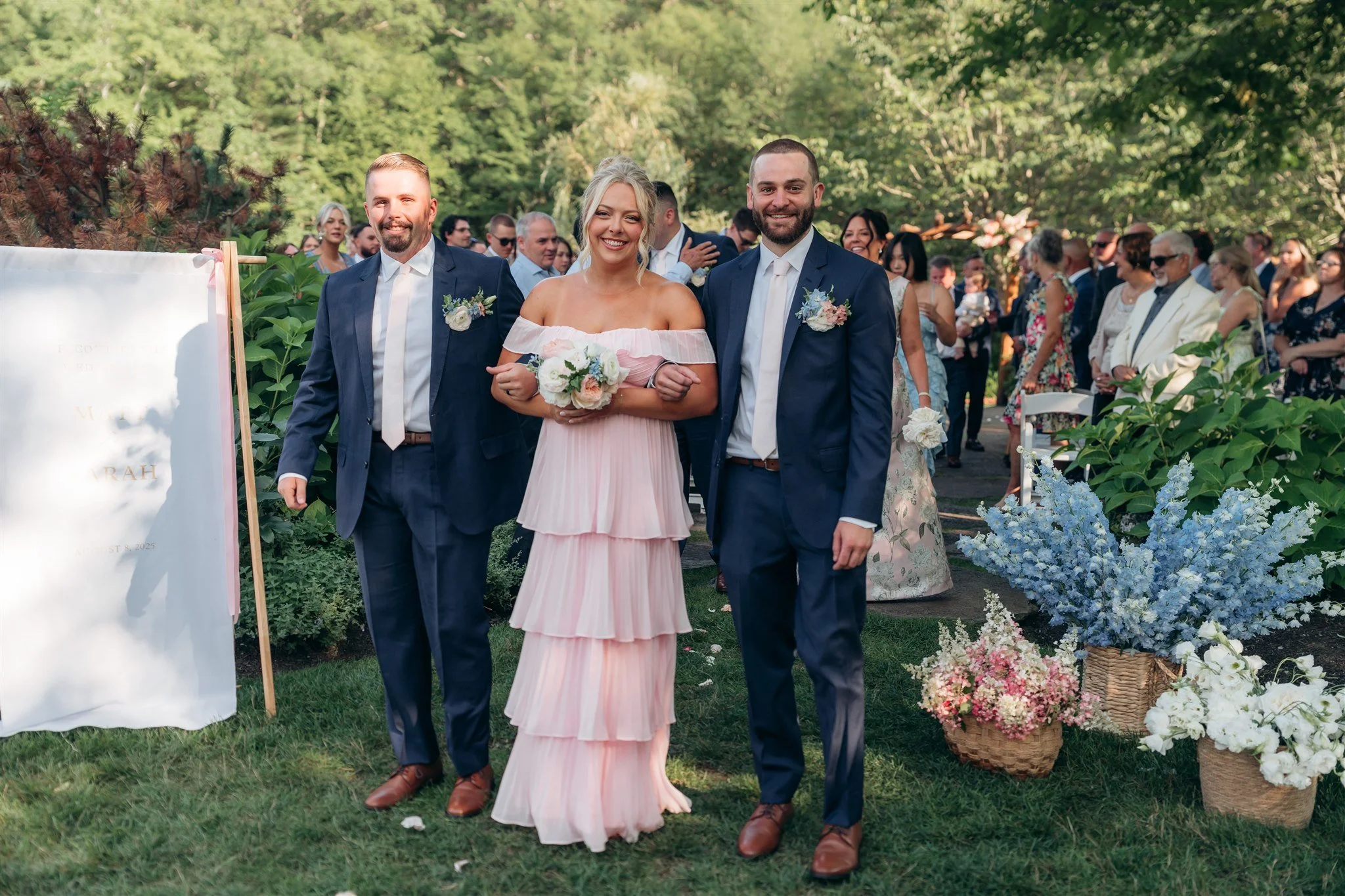 Bride walking down the aisle with two men, surrounded by wedding guests and decorated with flowers in an outdoor garden setting.