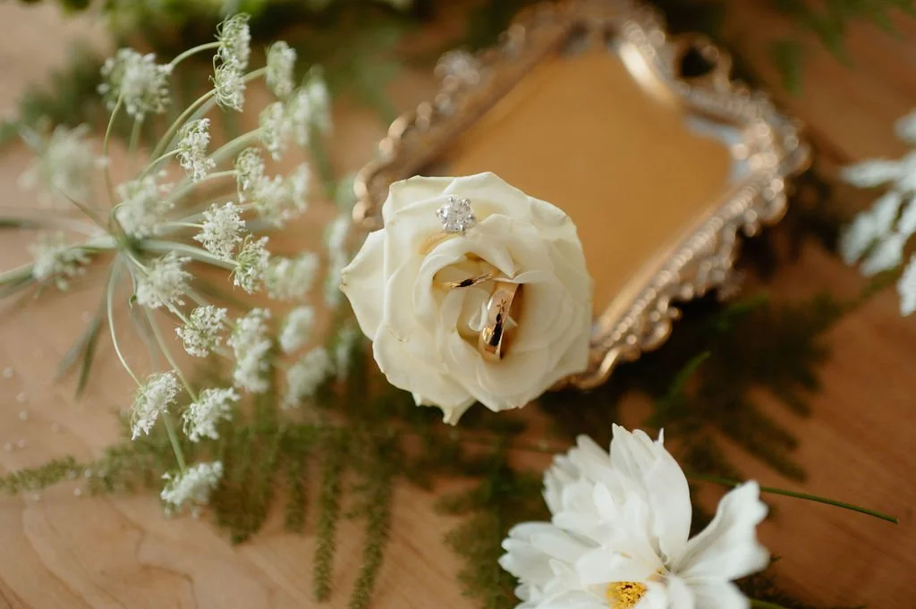 Wedding rings resting on a white rose with an ornate mirror and white flowers surrounding them.