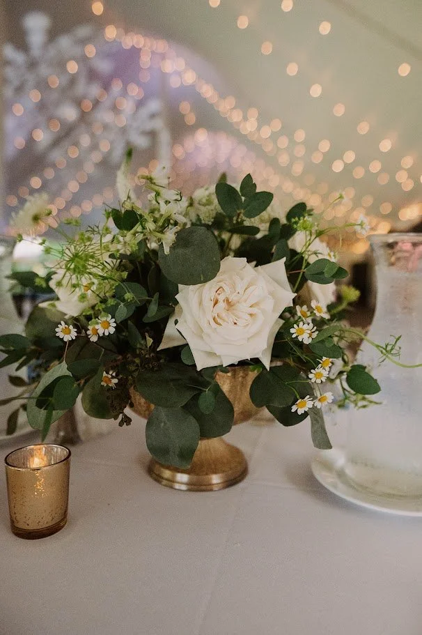 A floral centerpiece with white roses, small daisies, and greenery in a gold vase, surrounded by a gold candle holder and a glass jar, with wedding or party string lights in the background.