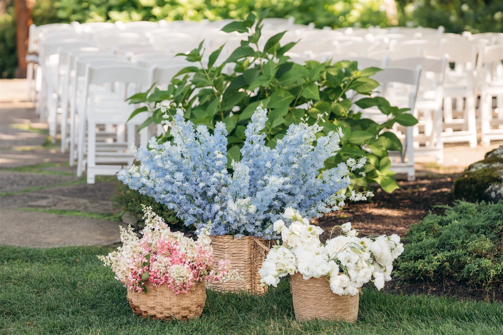 Three baskets filled with pink, blue, and white flowers sit on grass in front of white chairs arranged for an outdoor event. Green plants and trees are in the background.