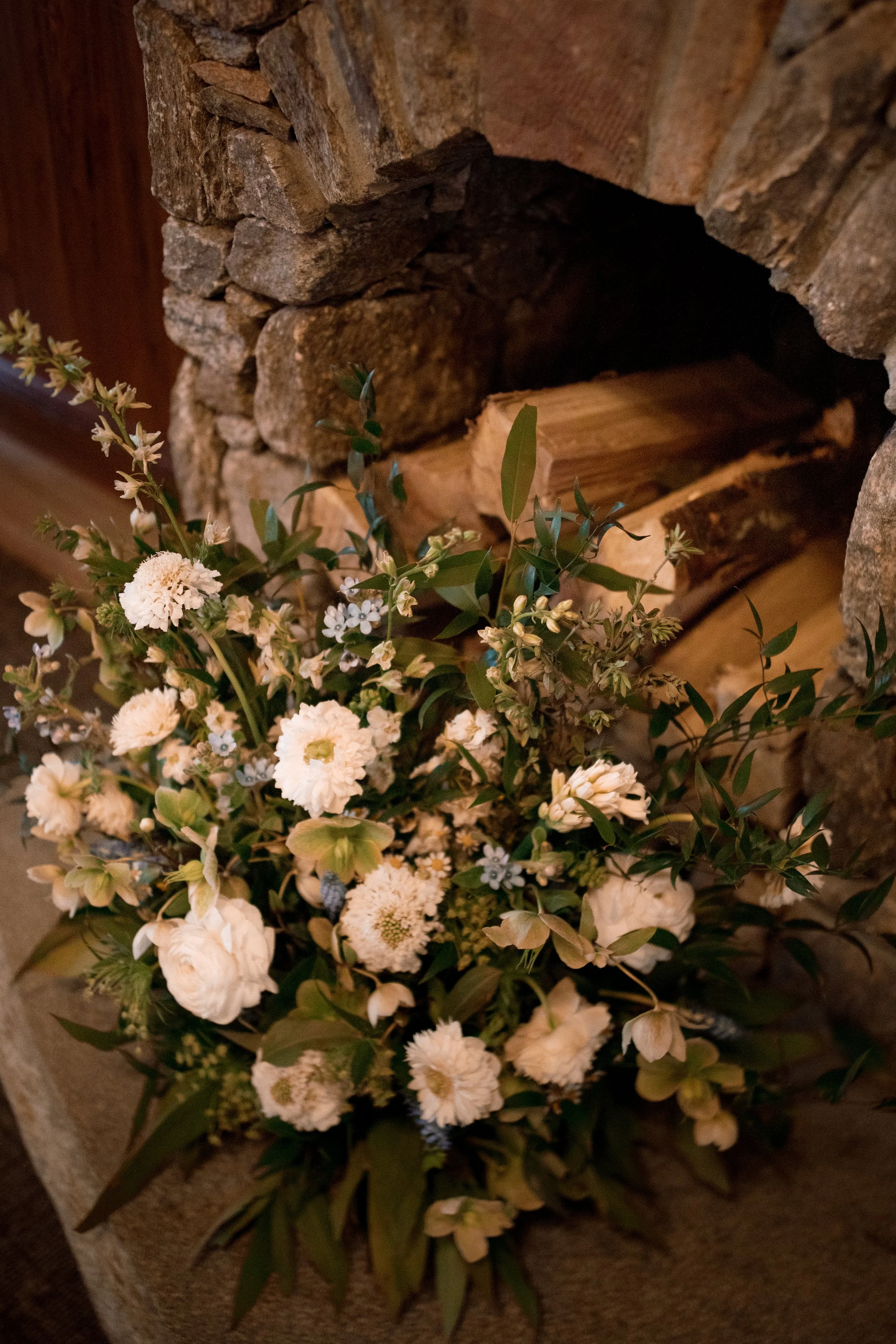A bouquet of white flowers placed in front of a rustic stone fireplace with firewood inside.