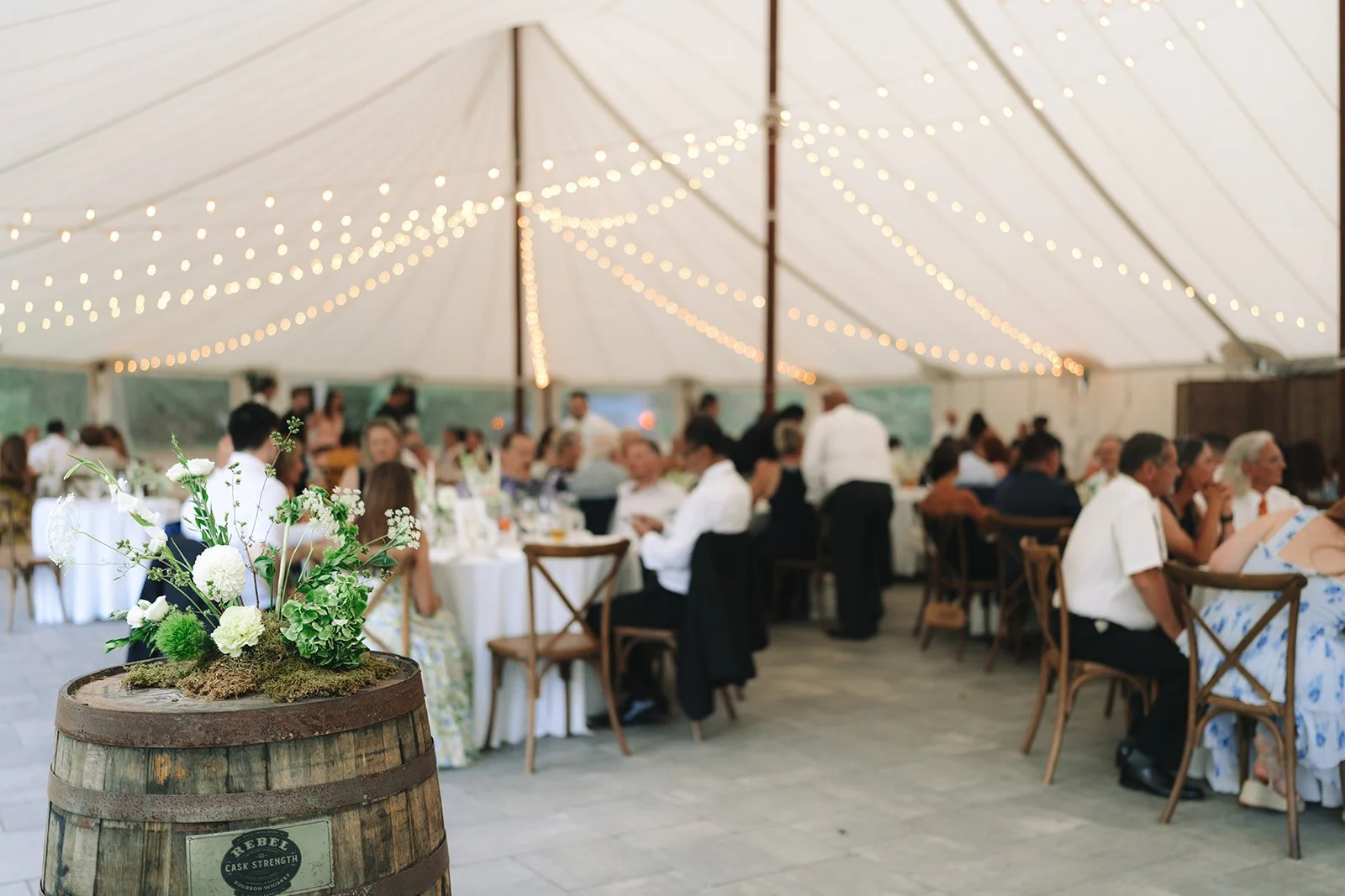 Decorative flower arrangement on a wooden barrel in front of a large white tent with string lights, hosting a wedding reception with guests seated at round and rectangular tables.