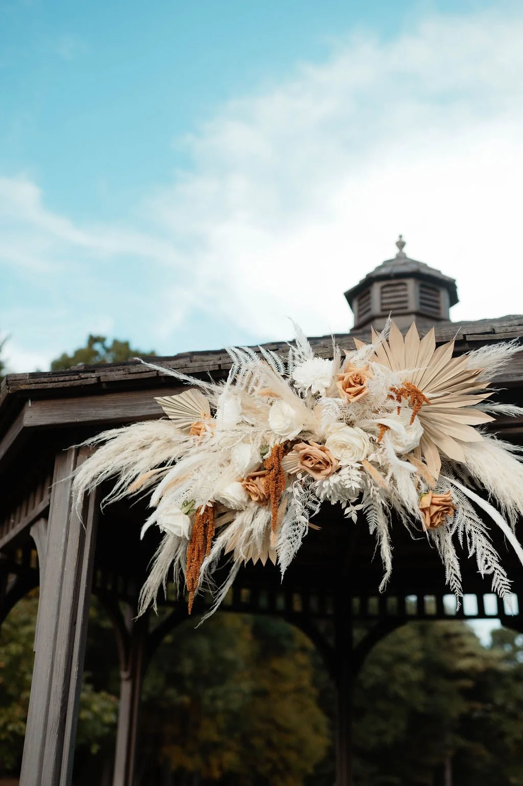 Decorative floral arrangement with white, beige, and brown flowers and dried foliage, attached to a wooden gazebo outdoors.