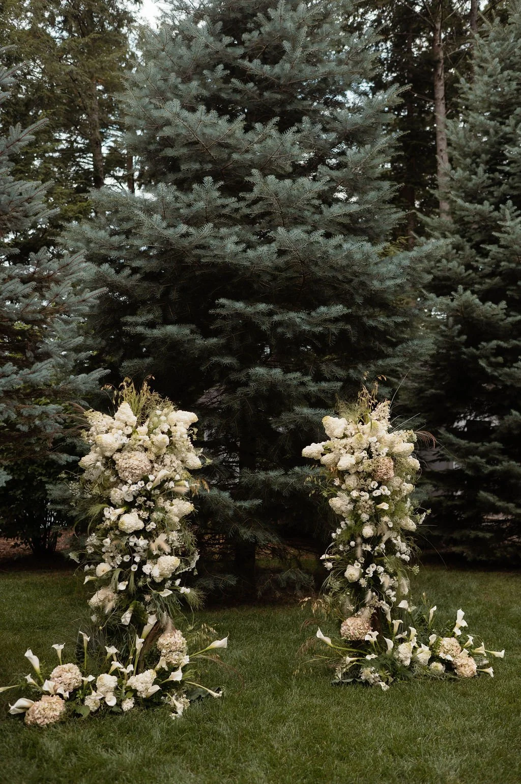 Two tall floral arrangements with white flowers and greenery, placed on grassy ground in front of a large evergreen tree.