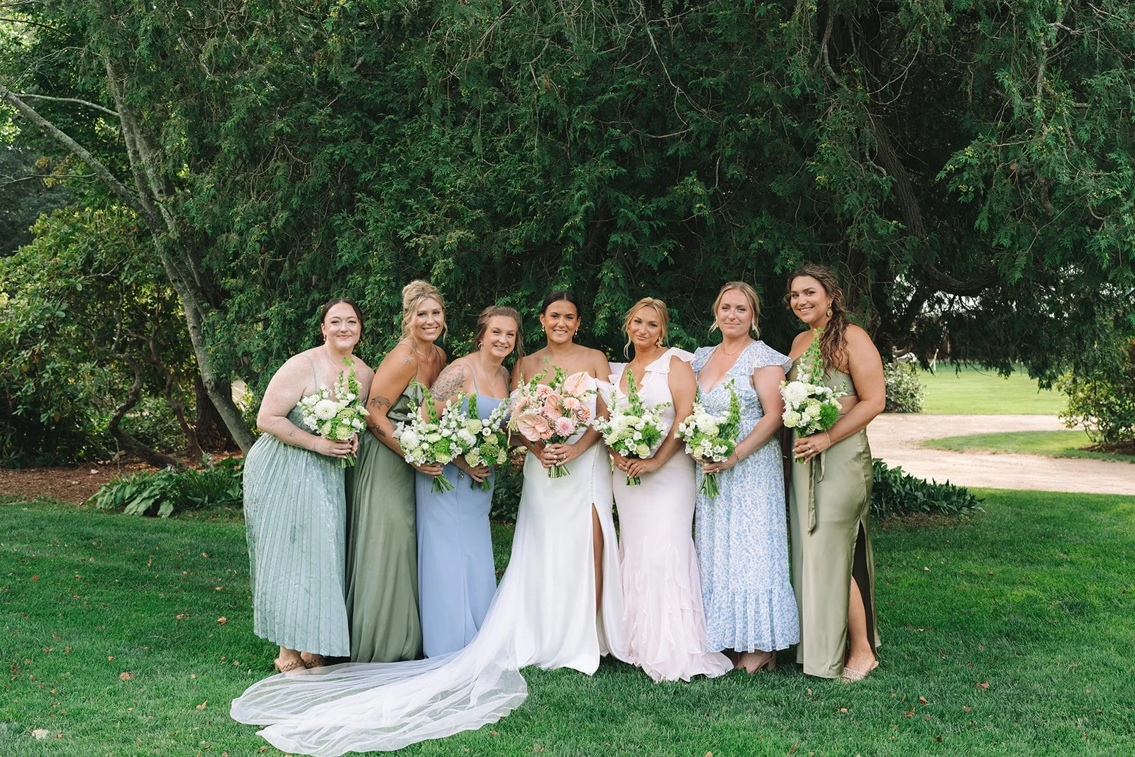 Group of women in pastel dresses holding white and pink flower bouquets, standing outdoors on grass with greenery and trees in the background at a wedding.