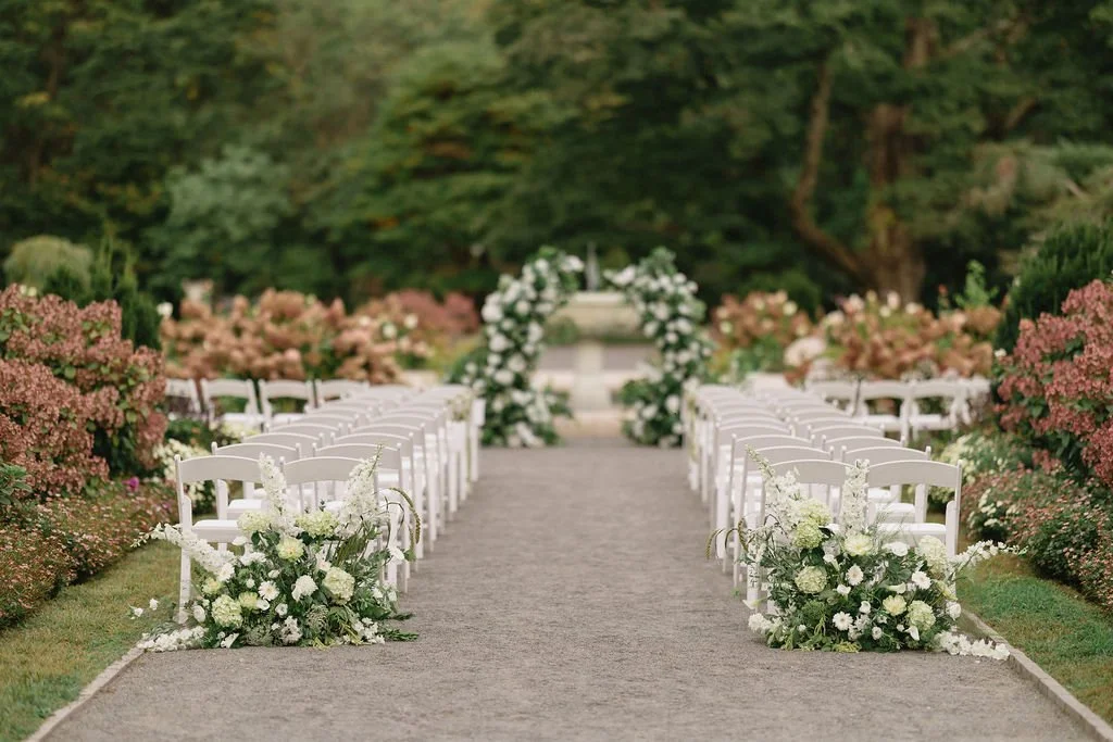 Outdoor wedding aisle decorated with white flowers and arranged chairs on both sides, leading to an arch with floral arrangements.