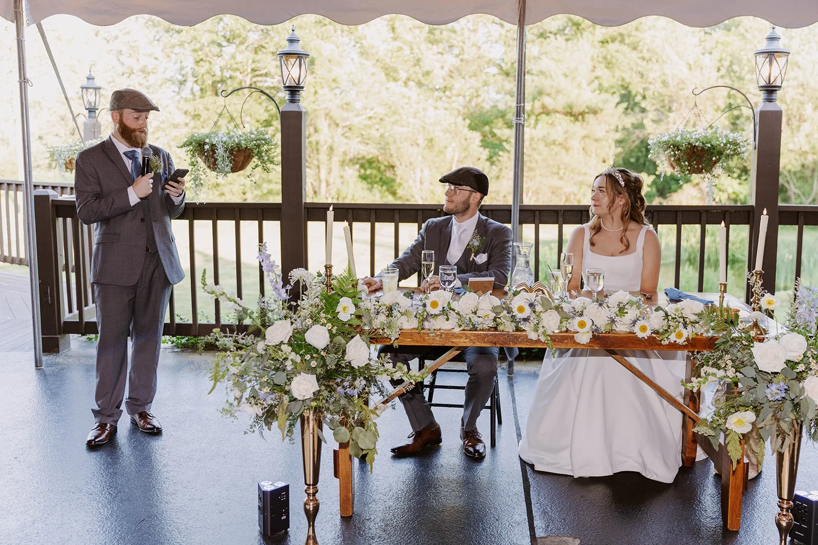 A wedding reception scene on an outdoor terrace with a man giving a speech while two people, a man and a woman, sit at a decorated table listening.