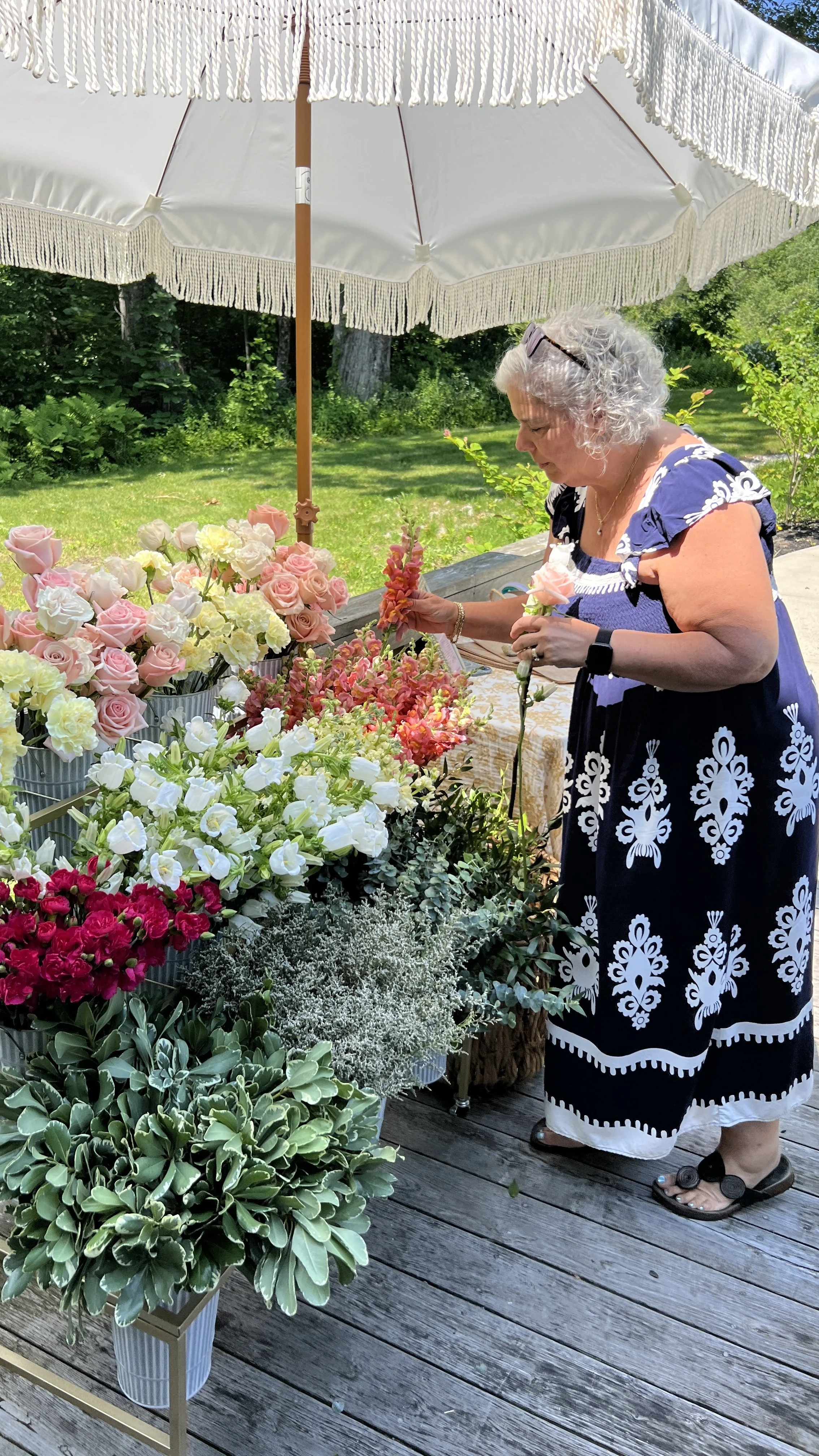 Older woman shopping for flowers at an outdoor market under a large white umbrella on a wooden deck surrounded by greenery.