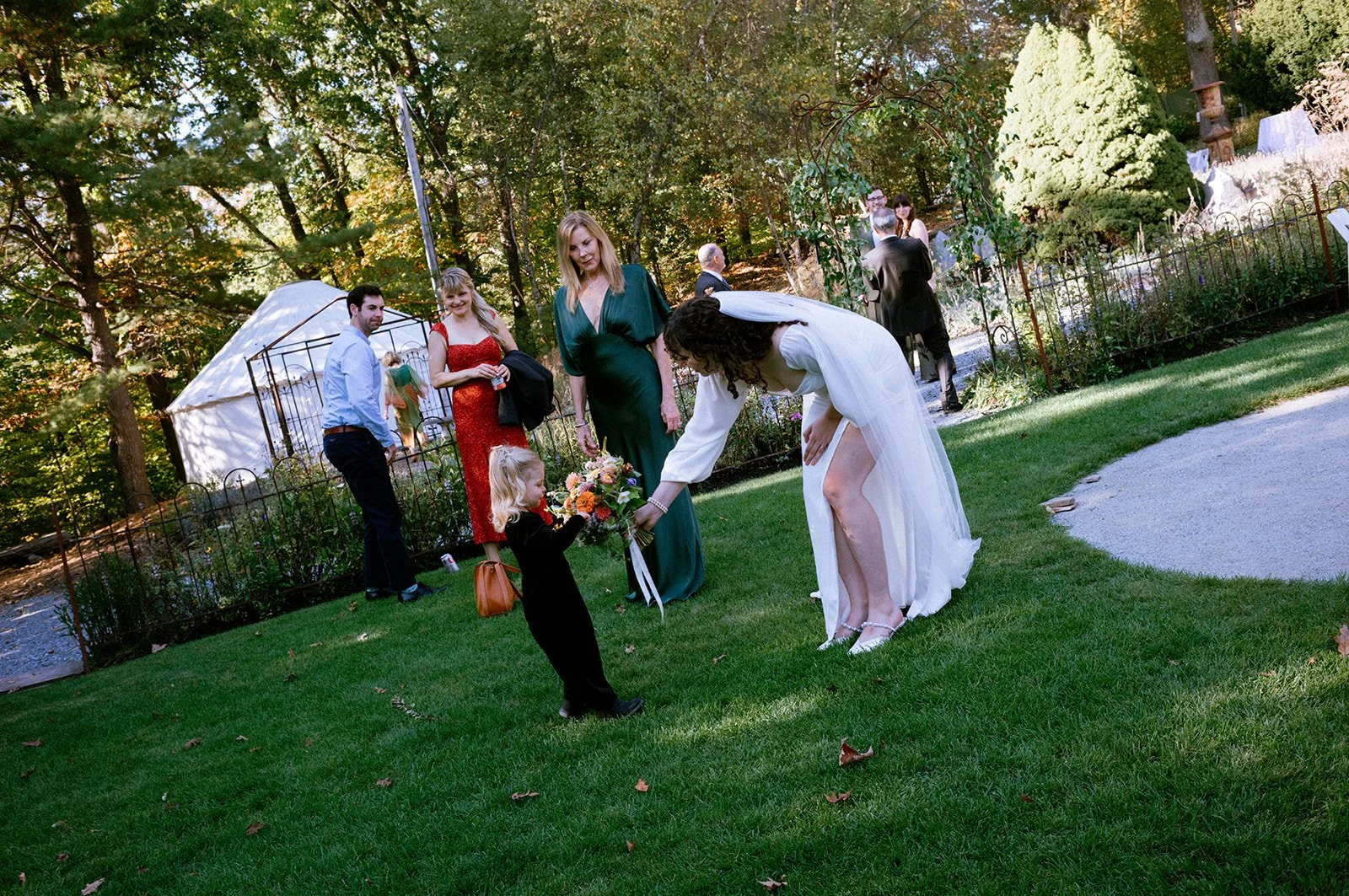 A woman in a white dress bending down to give flowers to a young girl in black clothing at an outdoor gathering, with other people in formal attire and trees in the background.