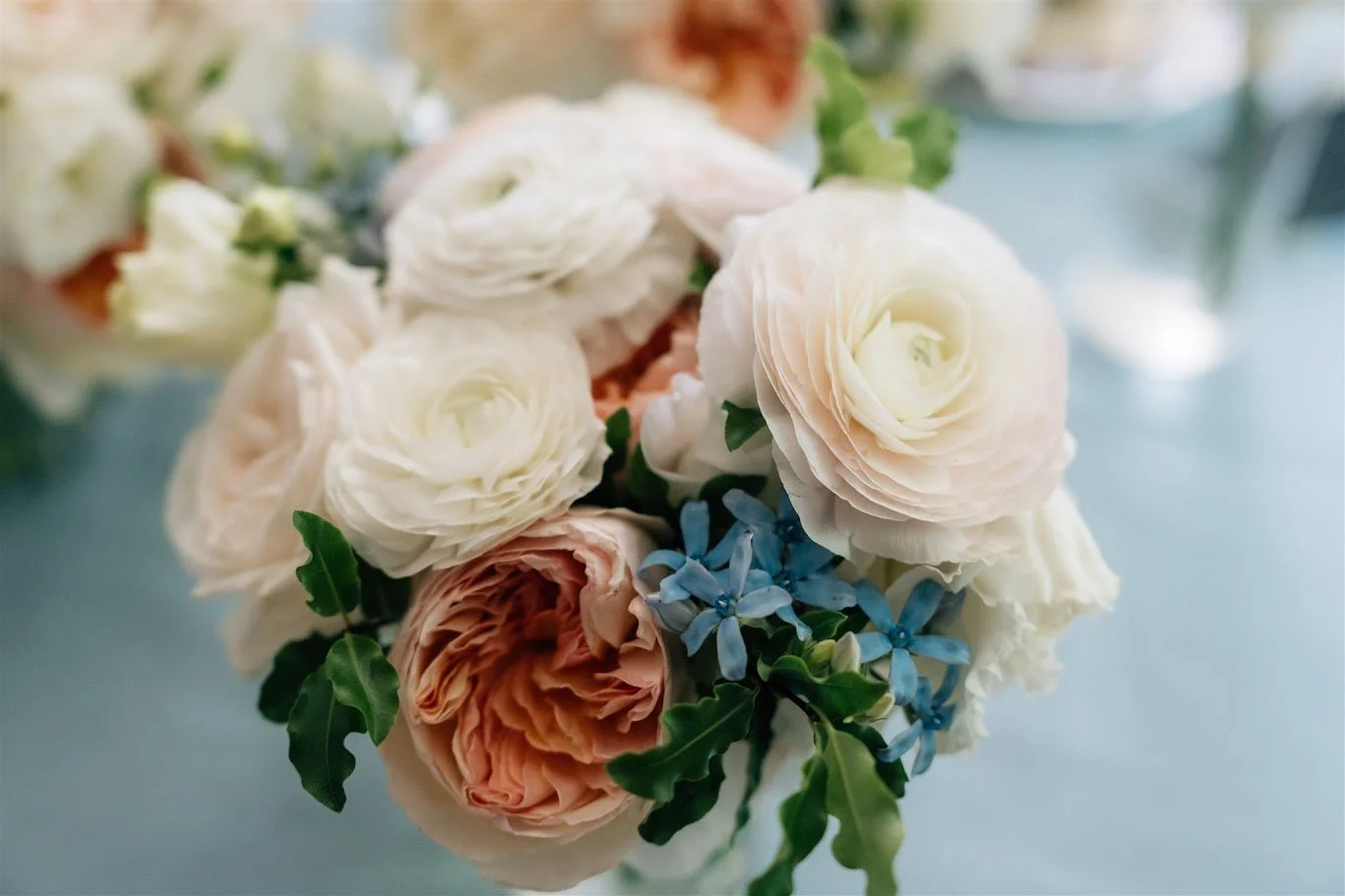 A flower arrangement in a white bowl with orange, yellow, pink, and white flowers on a wooden surface against a white textured wall.