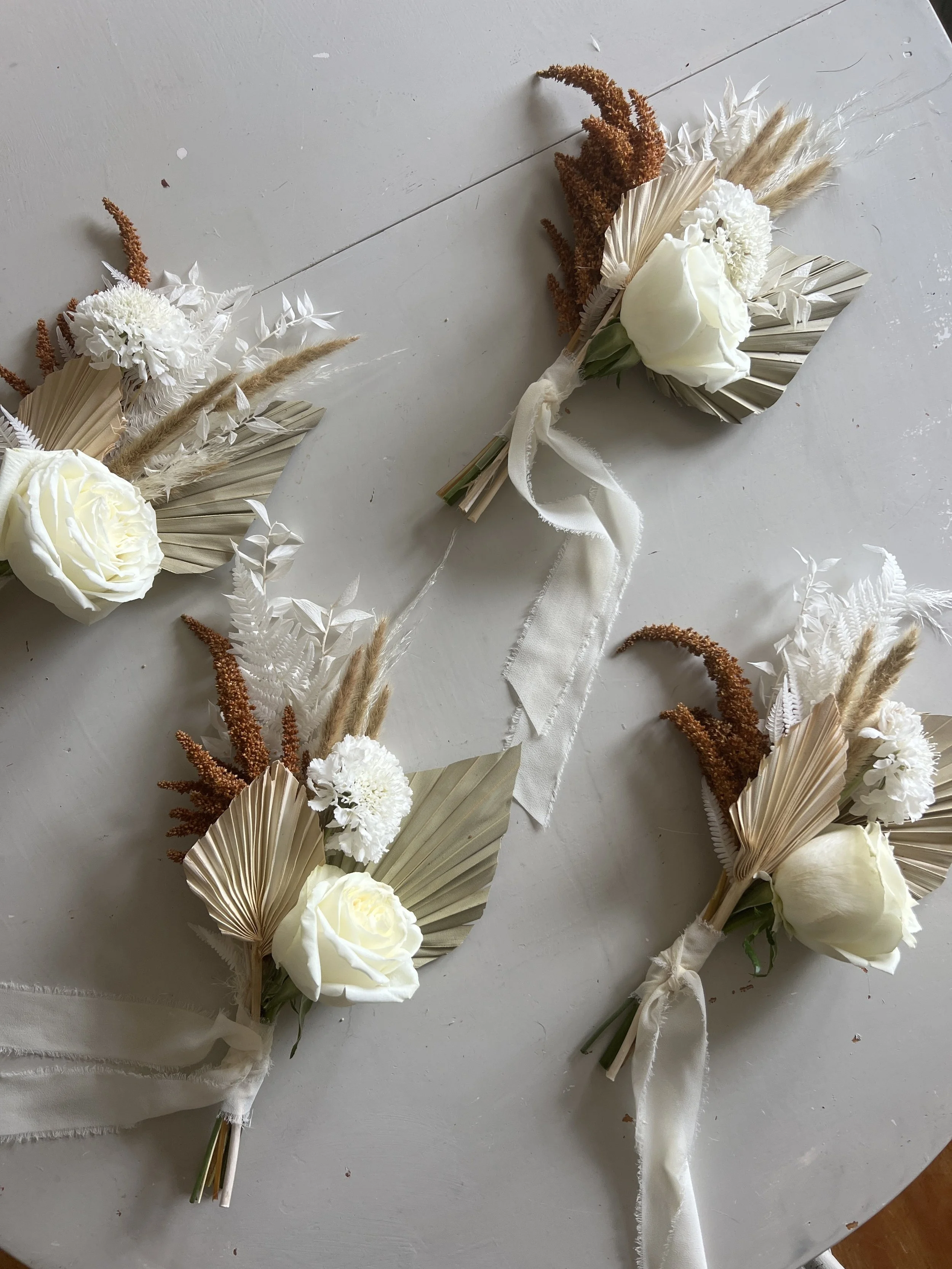 Three floral corsages with white roses, dried beige and rust-colored flowers, and decorative dried leaves on a gray surface.