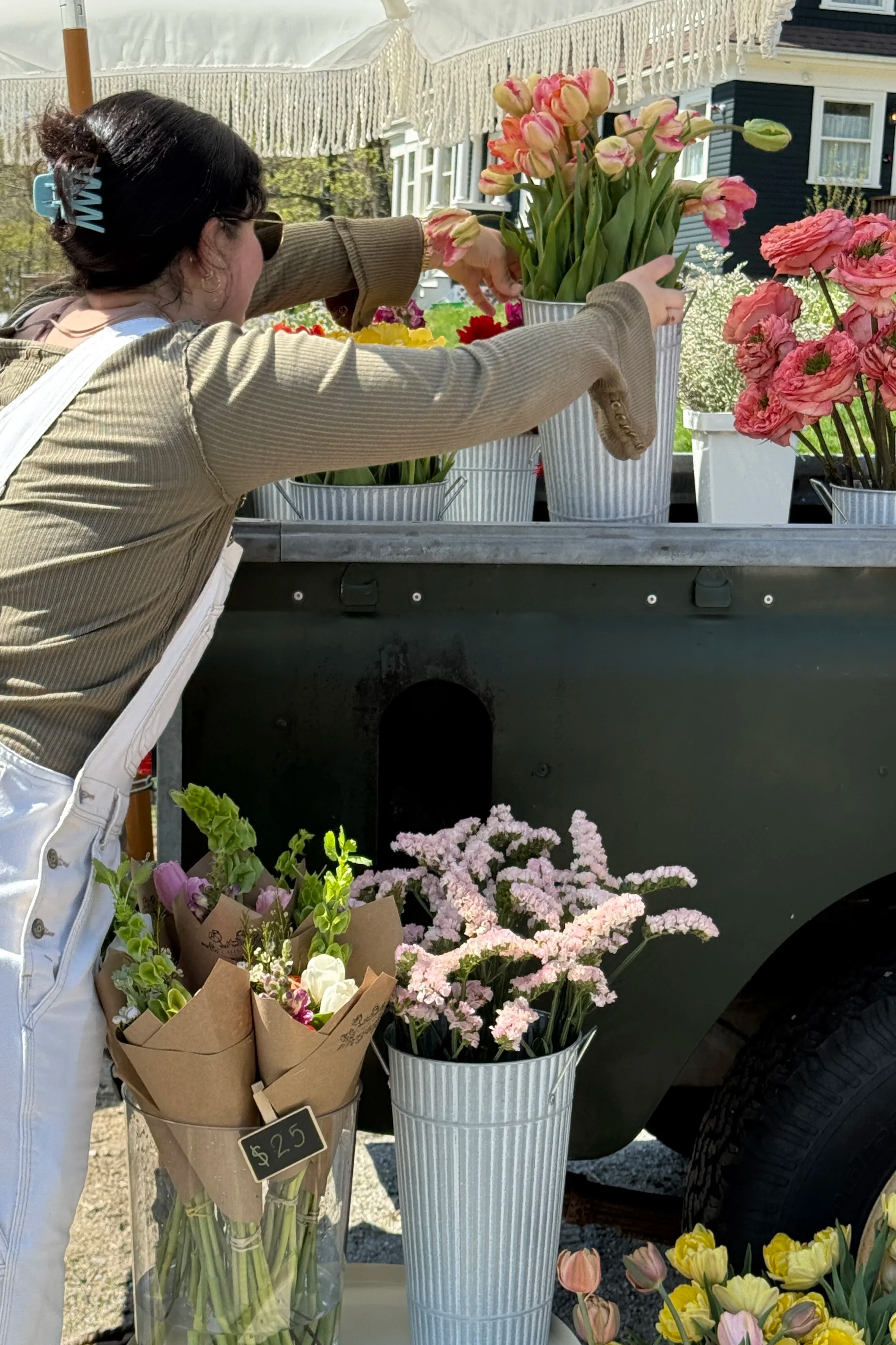 A woman arranging pink tulips in a white ceramic pot at a flower stand. The stand has other flowers like pink, yellow, and purple blooms, with a clear glass jar containing purple and white flowers with a $25 price tag. The scene is outdoors, with a house visible in the background.