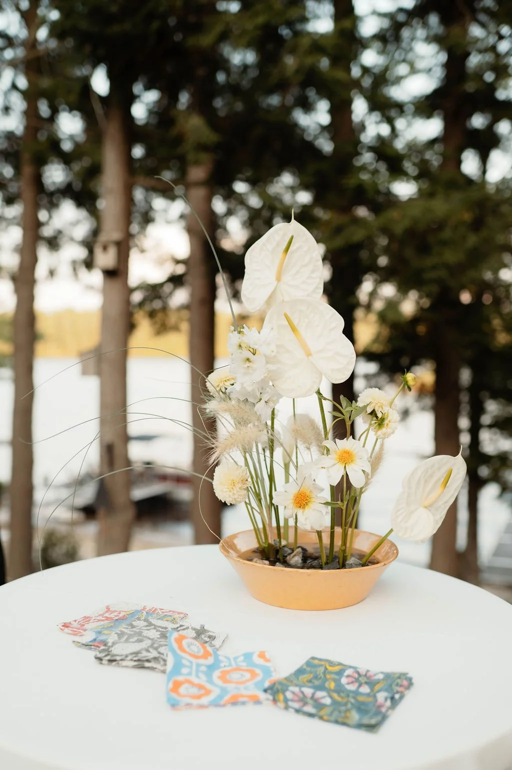A floral arrangement with white flowers and anthuriums in an orange bowl, placed on a white table outdoors, with patterned fabric pieces on the table and trees in the background.